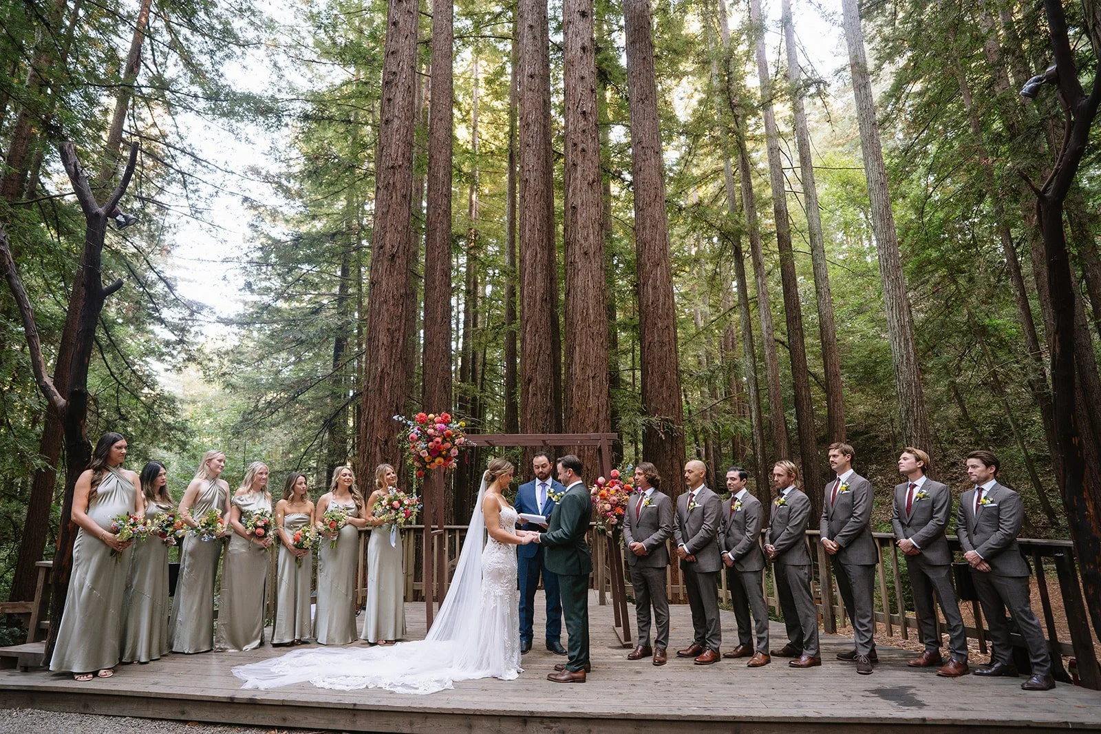 A wedding ceremony taking place outdoors in a forest with tall trees. The bride and groom are in the center, exchanging vows, with the officiant standing behind them. The bride is wearing a white lace gown with a long train, and the groom is in a dark suit. Bridesmaids in matching silver dresses and groomsmen in gray suits with maroon ties stand on either side.
