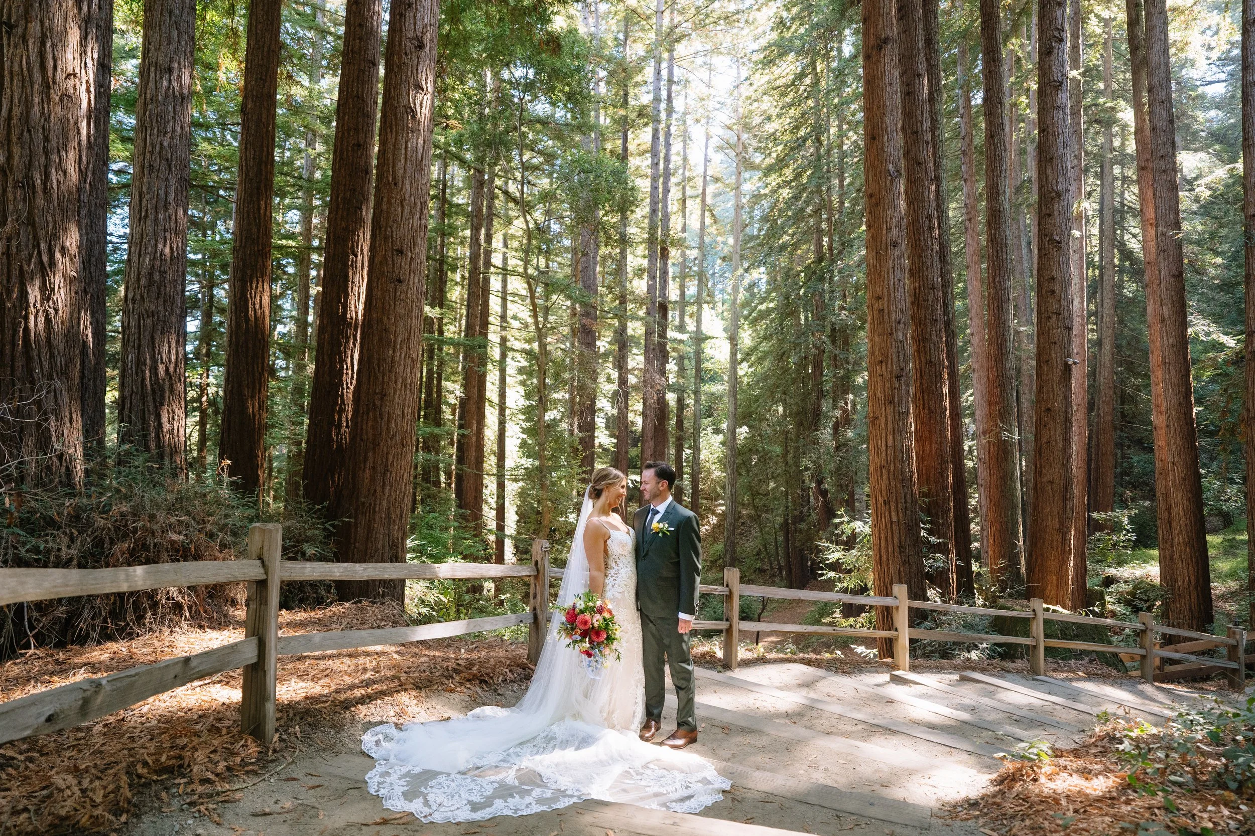 A bride and groom stand close together in a wooded forest, holding a bouquet of flowers, during their wedding photo session.