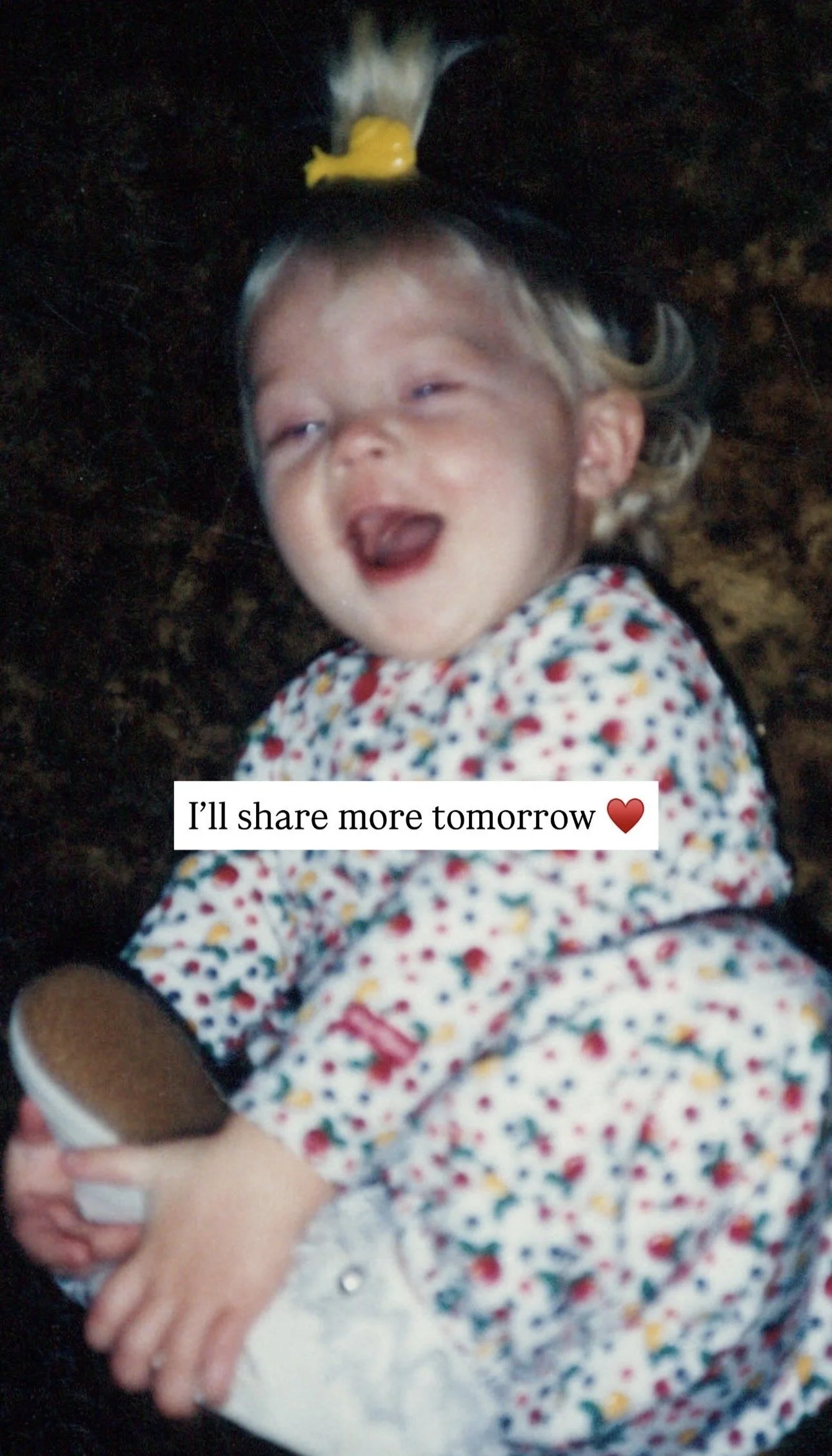 A young girl with a yellow bow in her hair, wearing a floral dress, holding a small container, smiling with eyes half closed, with text overlay 'I'll share more tomorrow' accompanied by a red heart emoji.