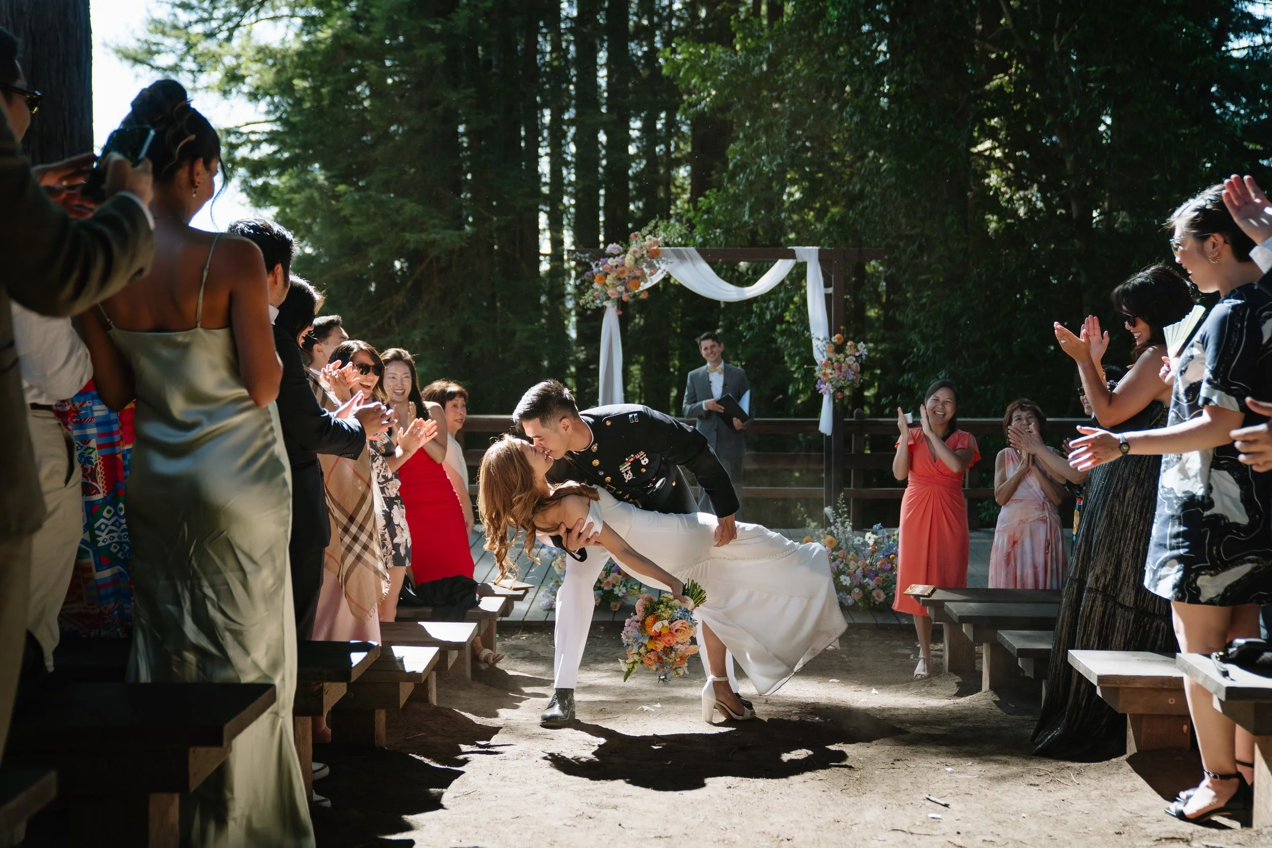 A wedding ceremony in an outdoor forest setting, with a couple kissing at the center while guests cheer and applaud.