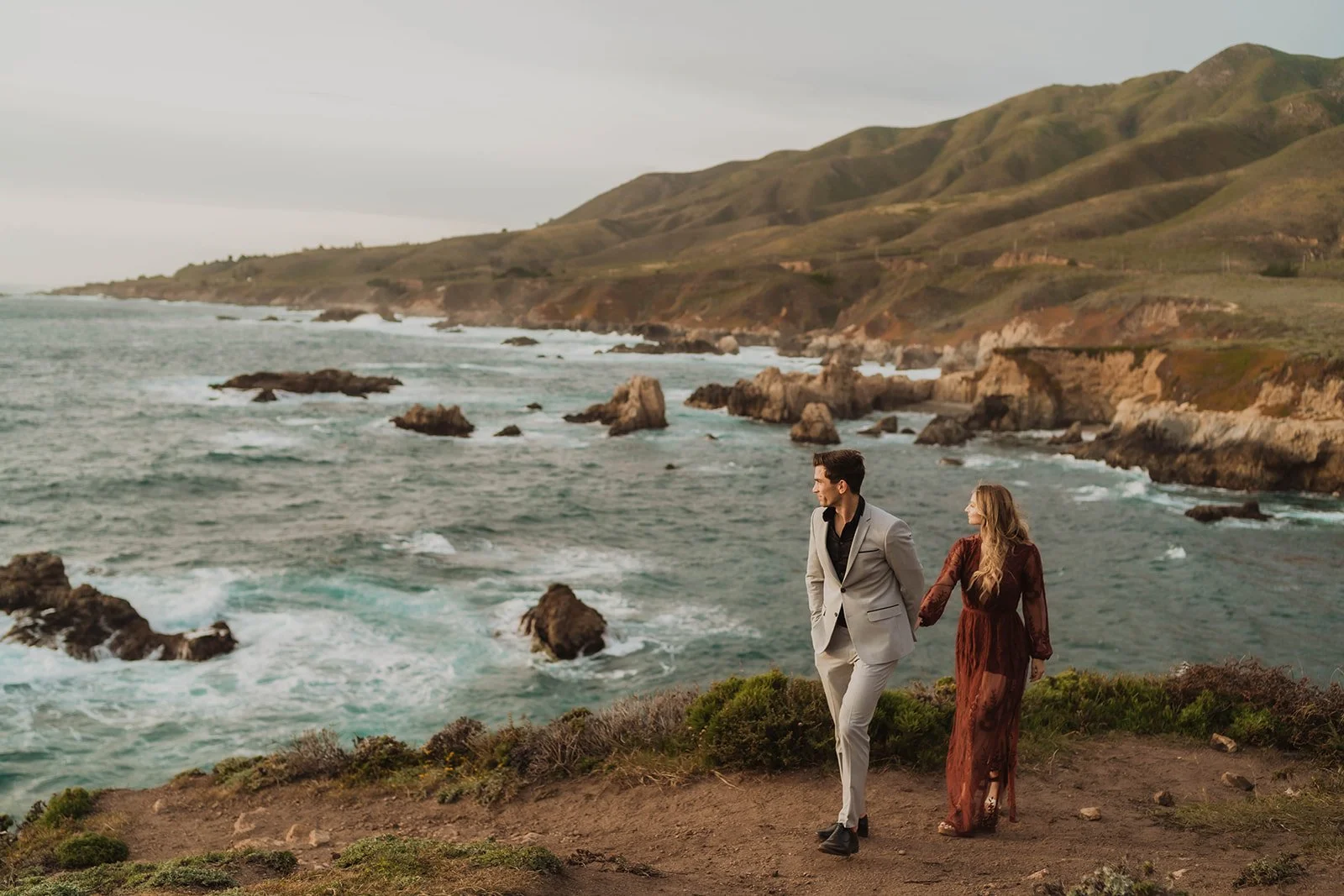 A man and woman holding hands walking along a coastal cliffside. The man is in a gray suit, and the woman is in a long, brown dress. The ocean with rocks and green hills are visible in the background.