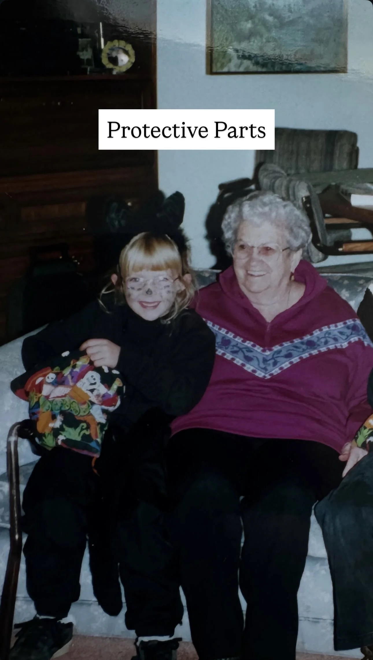 A young girl wearing black clothes and face paint sitting next to an elderly woman with gray hair, both smiling, in a living room. A sign that reads 'Protective Parts' is above them.
