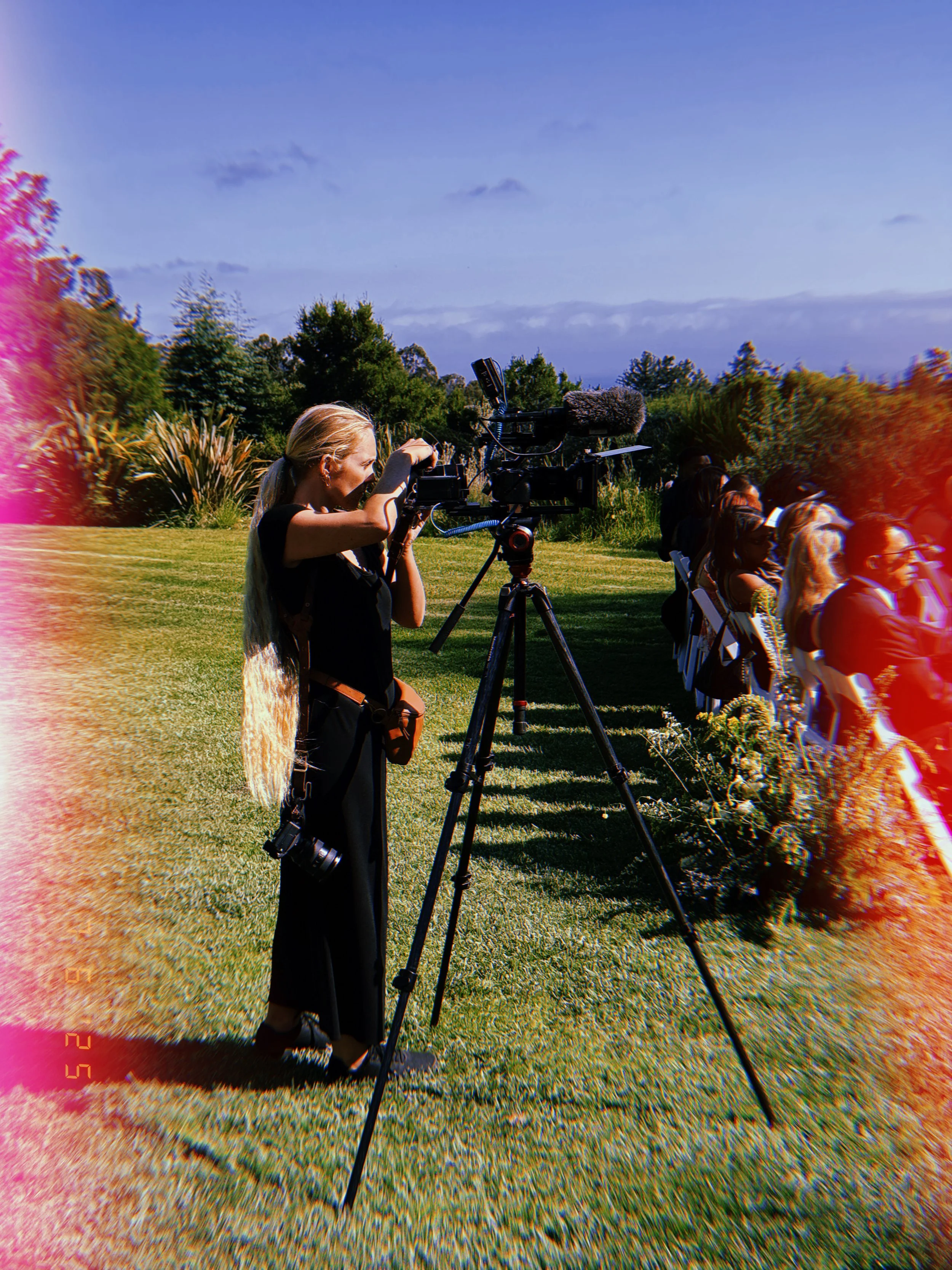 A woman filming with a professional camera on a tripod at an outdoor event, with people seated on chairs beside a landscaped garden under a partly cloudy sky.