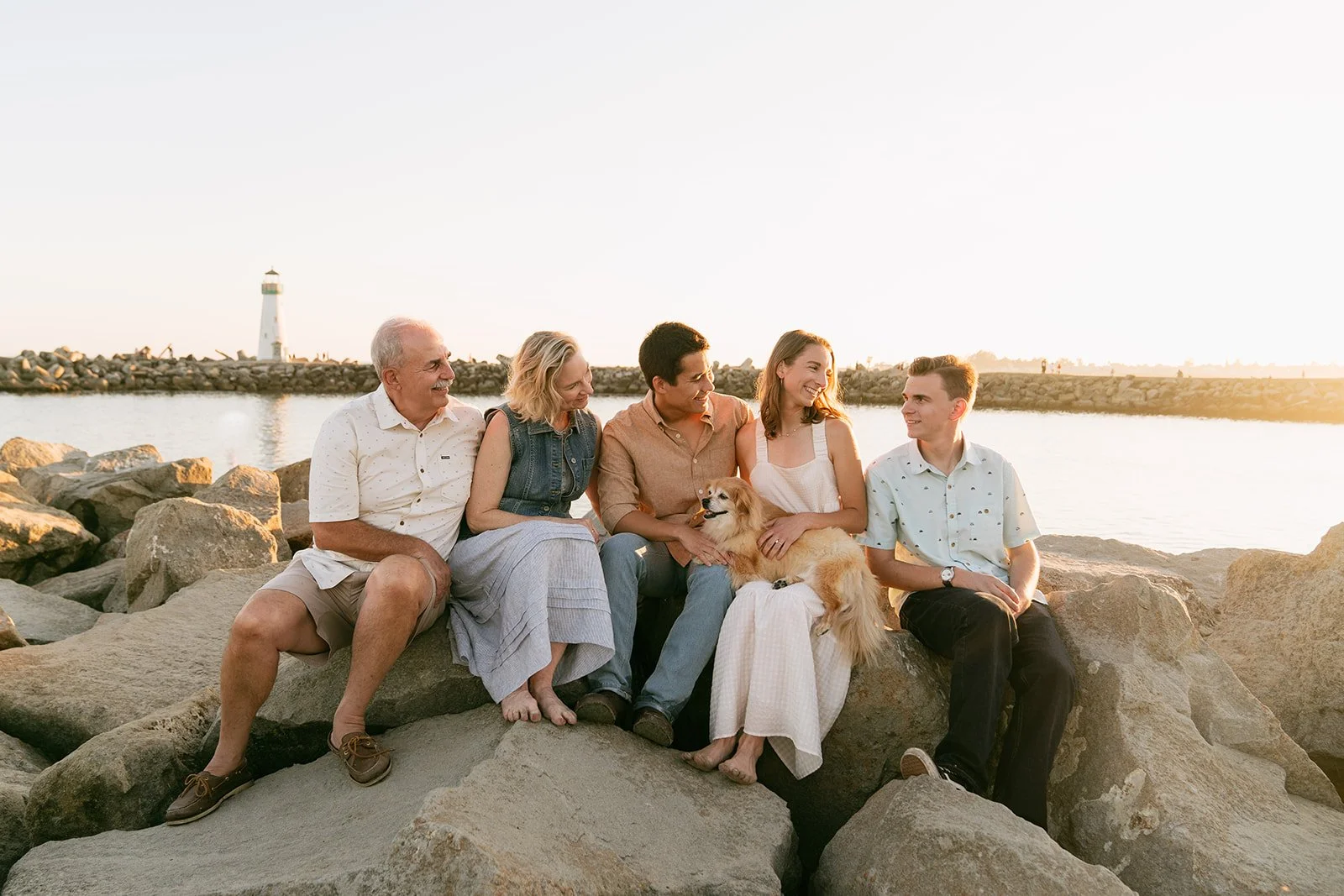 A multi-generational family sitting on rocks by the water at sunset, smiling and enjoying each other's company, with a lighthouse in the background.
