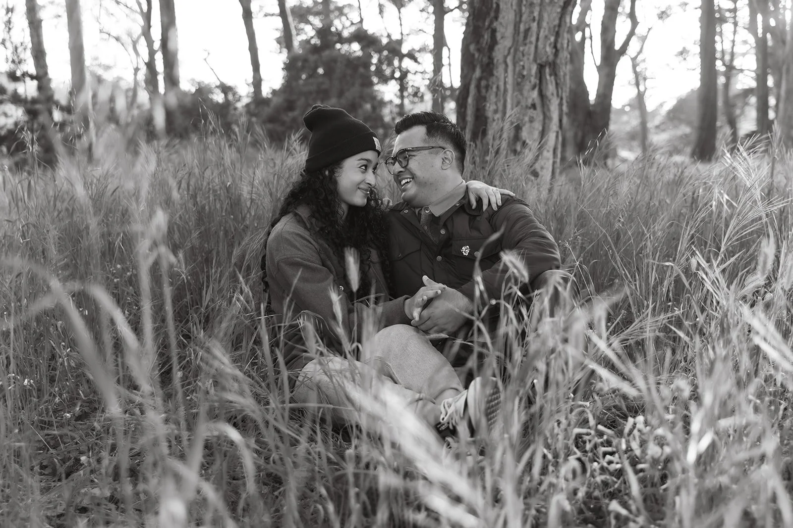 A young couple sitting close together in tall grass, smiling and holding hands, in a wooded area with trees in the background.