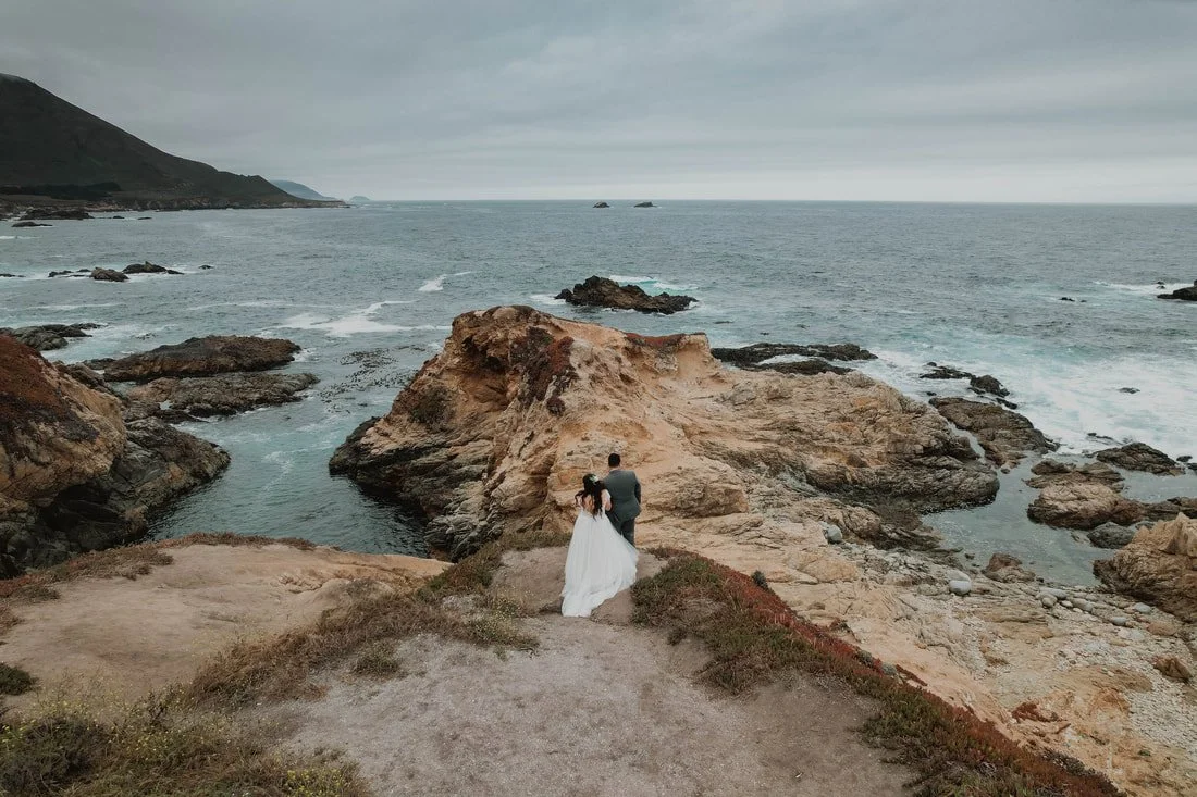A bride and groom standing on rocky cliffs overlooking the ocean, with a cloudy sky above.