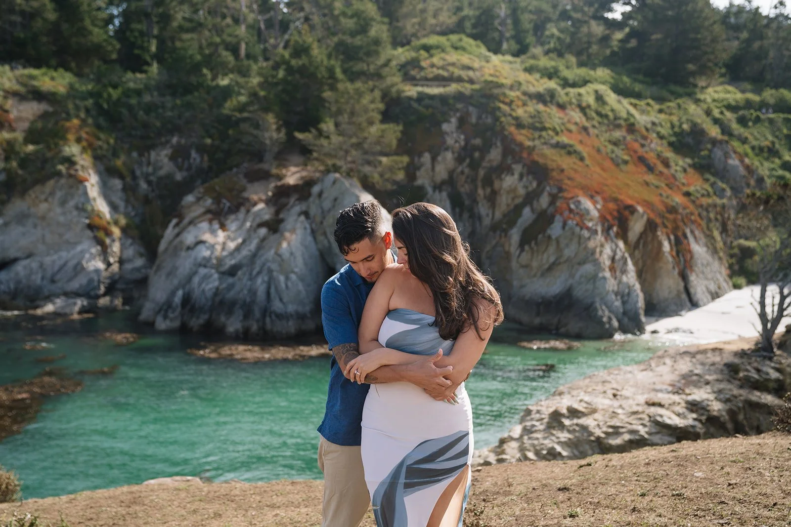 A couple hugging near a river with rocky cliffs and green foliage in the background.