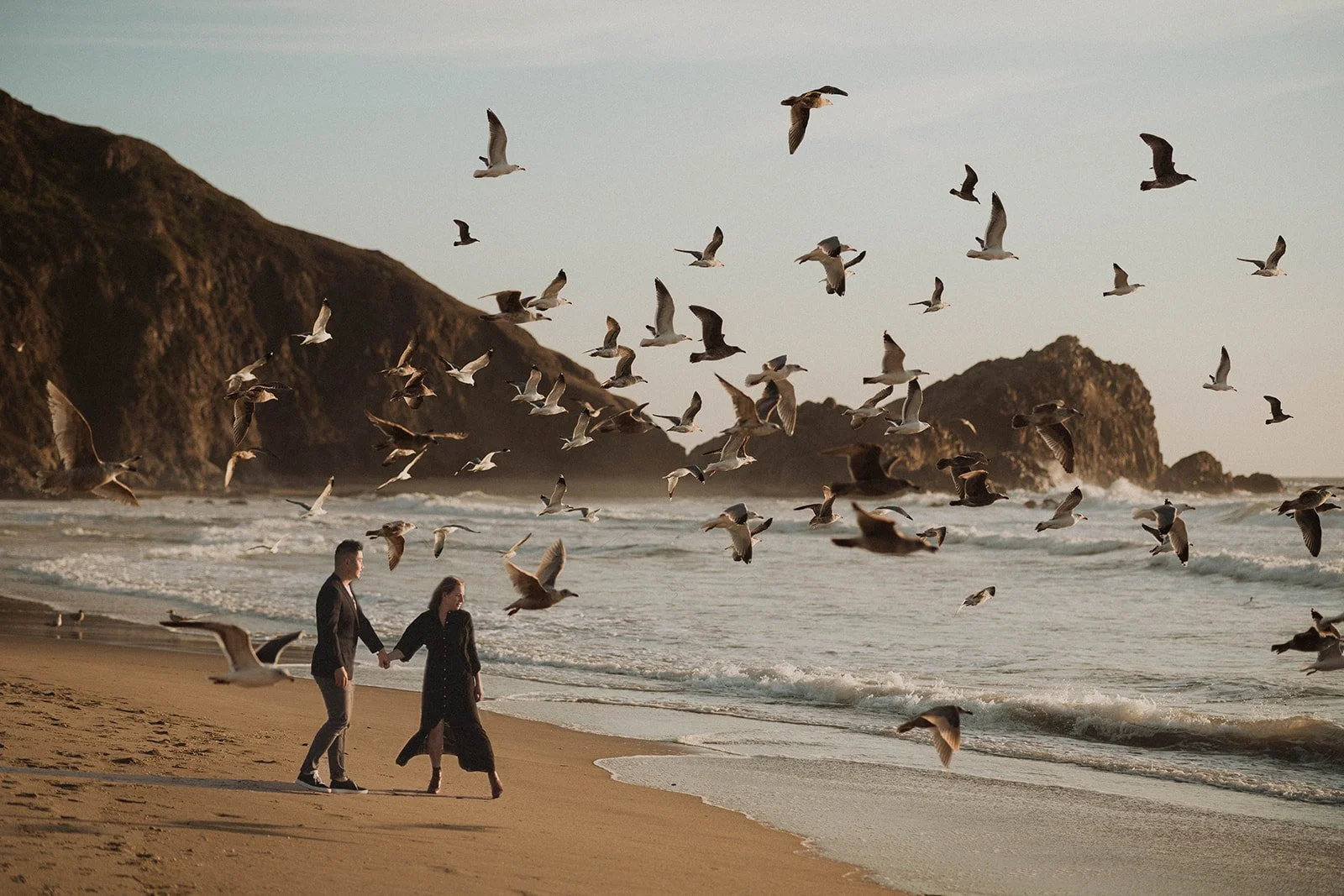 A couple walking hand in hand on a beach with seagulls flying overhead and rocky cliffs in the background.