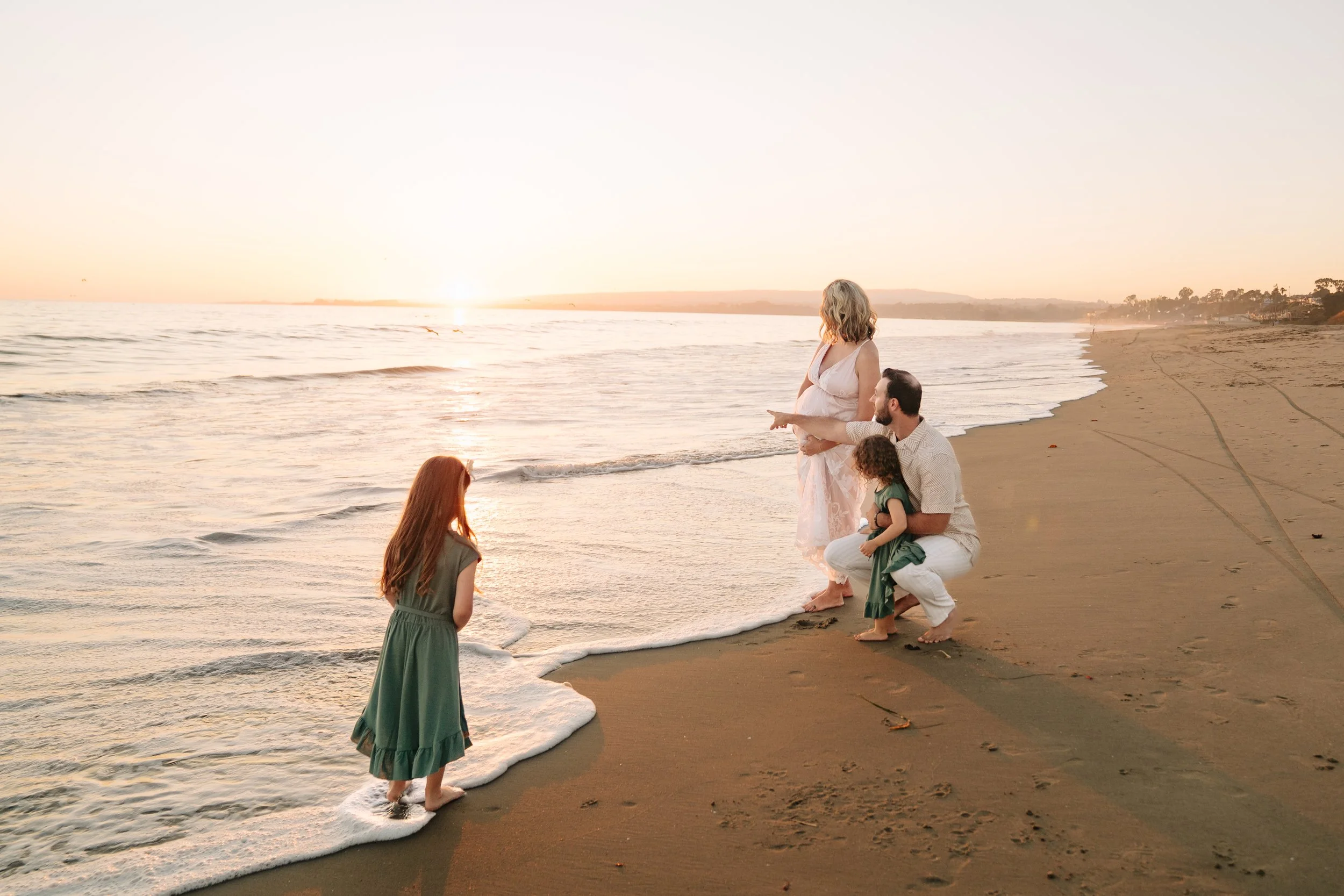 A family of four on a beach at sunset, with the mother holding her pregnant belly, the father kneeling, and two young girls standing in the sand near the water.