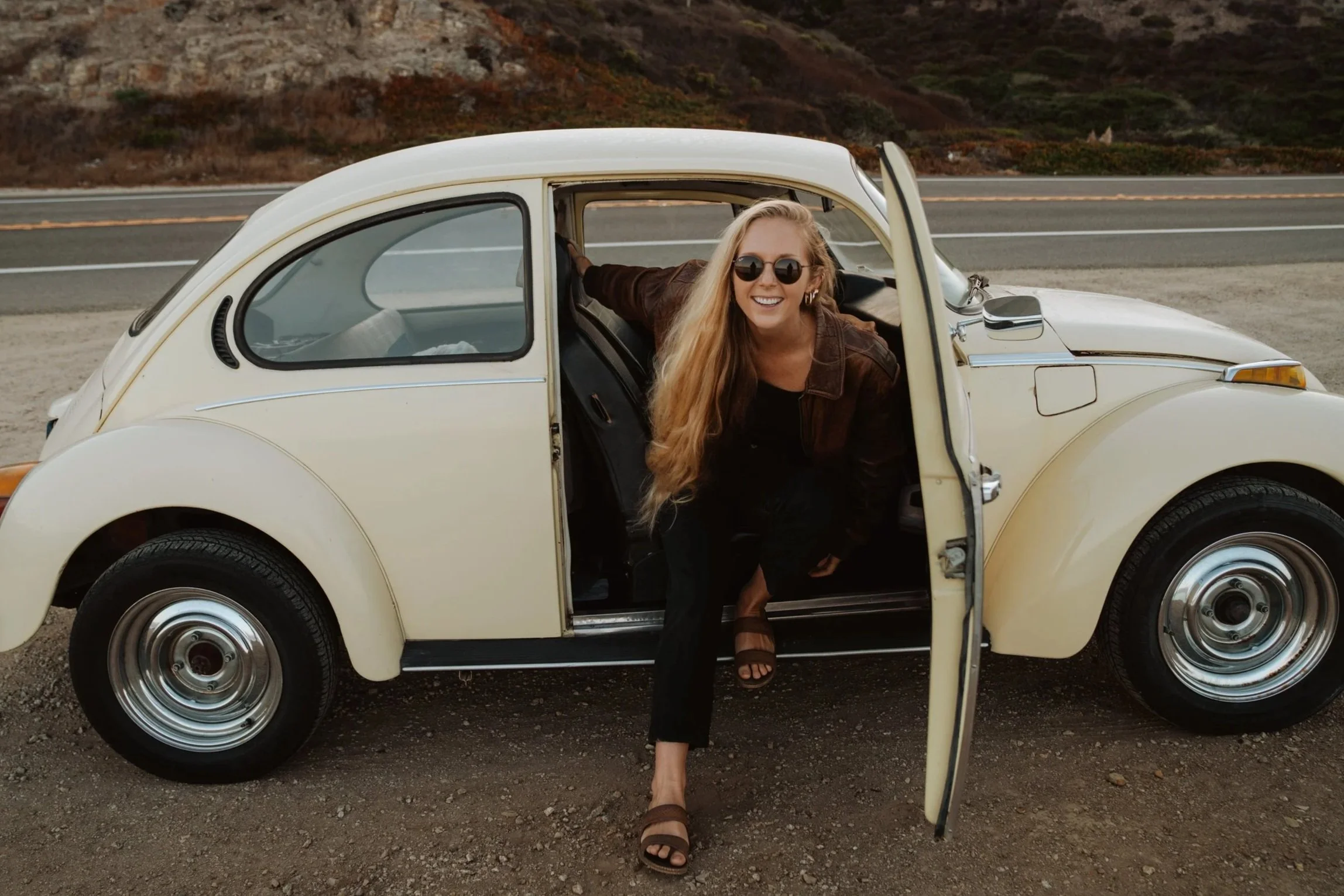 Woman with long blonde hair, wearing sunglasses and a brown jacket, smiling and leaning out of the open door of a vintage cream-colored Volkswagen Beetle parked on the side of a rural highway with hills in the background.