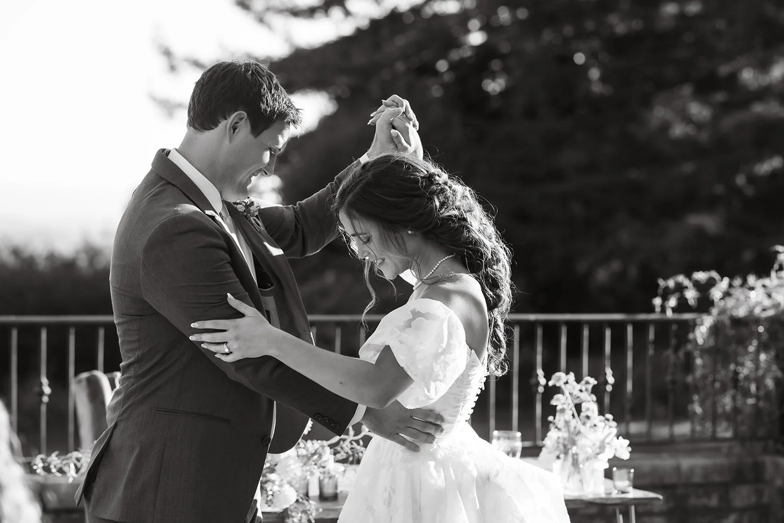 A black and white photo of a bride and groom dancing at their wedding, smiling and holding hands.