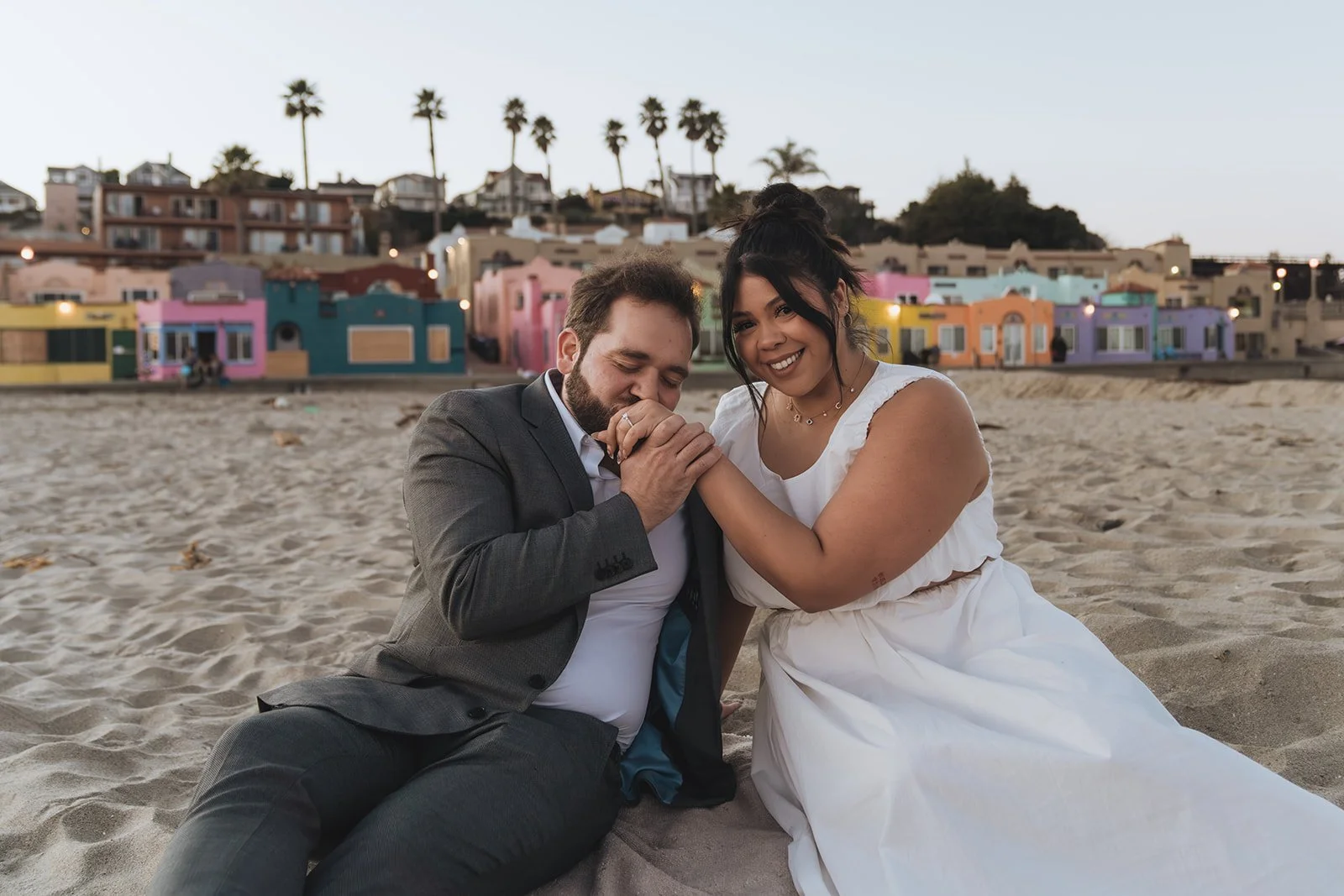 A couple, a man in a suit and a woman in a white dress, sitting on the beach at sunset, holding hands and smiling with colorful houses and palm trees in the background.