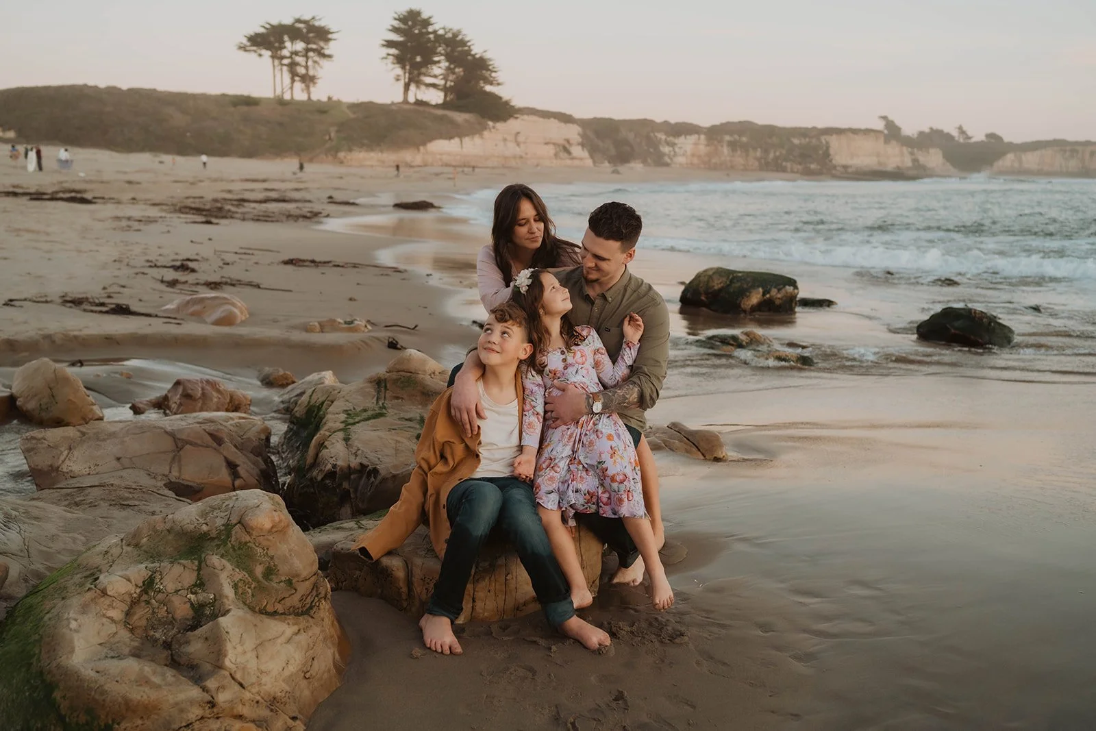 A family of four sitting on rocks at the beach during sunset, with waves, cliffs, and trees in the background.