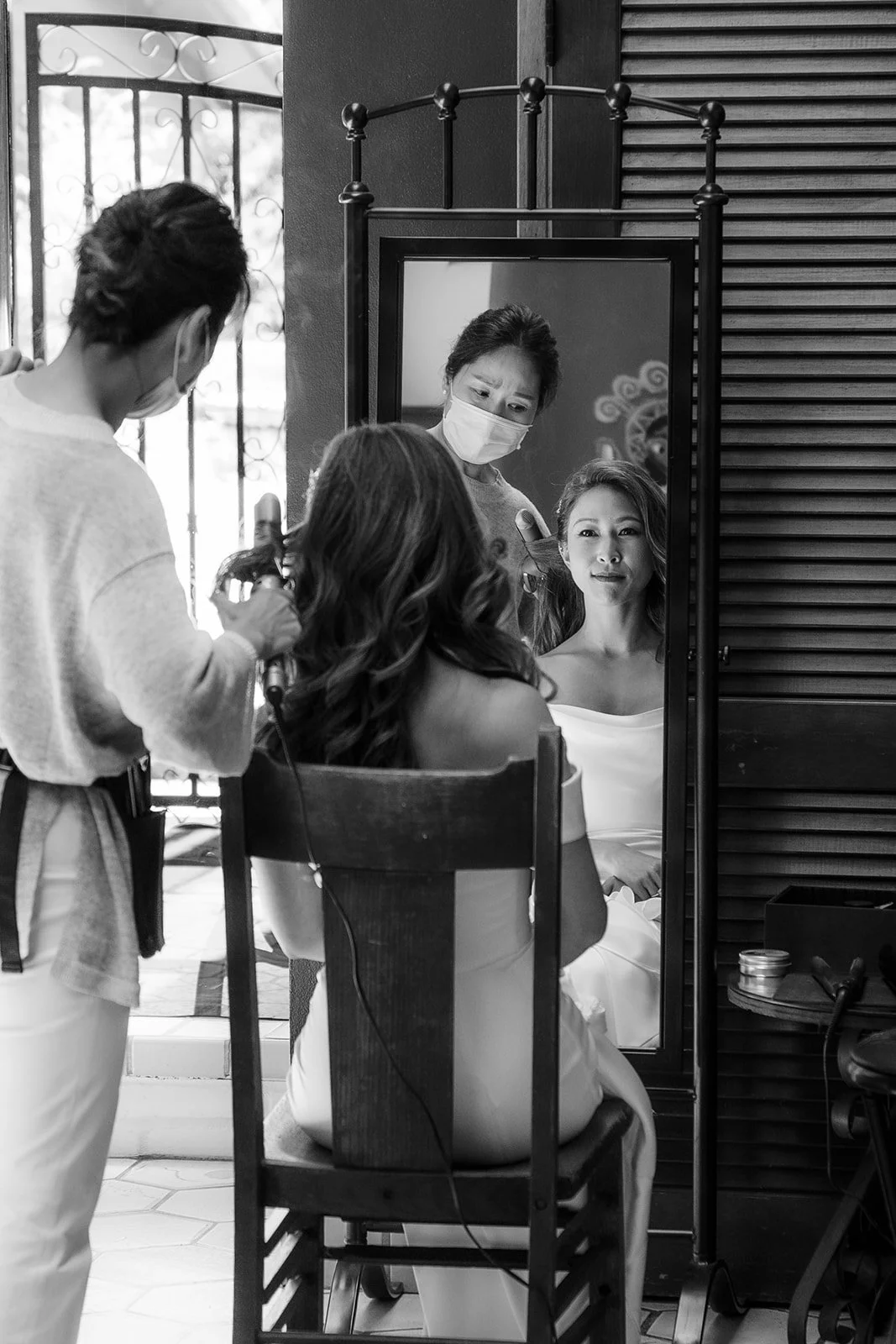 A woman with curled hair is sitting in a chair and having her hair done by a hairstylist, while looking at her reflection in a mirror. Both women are wearing masks.