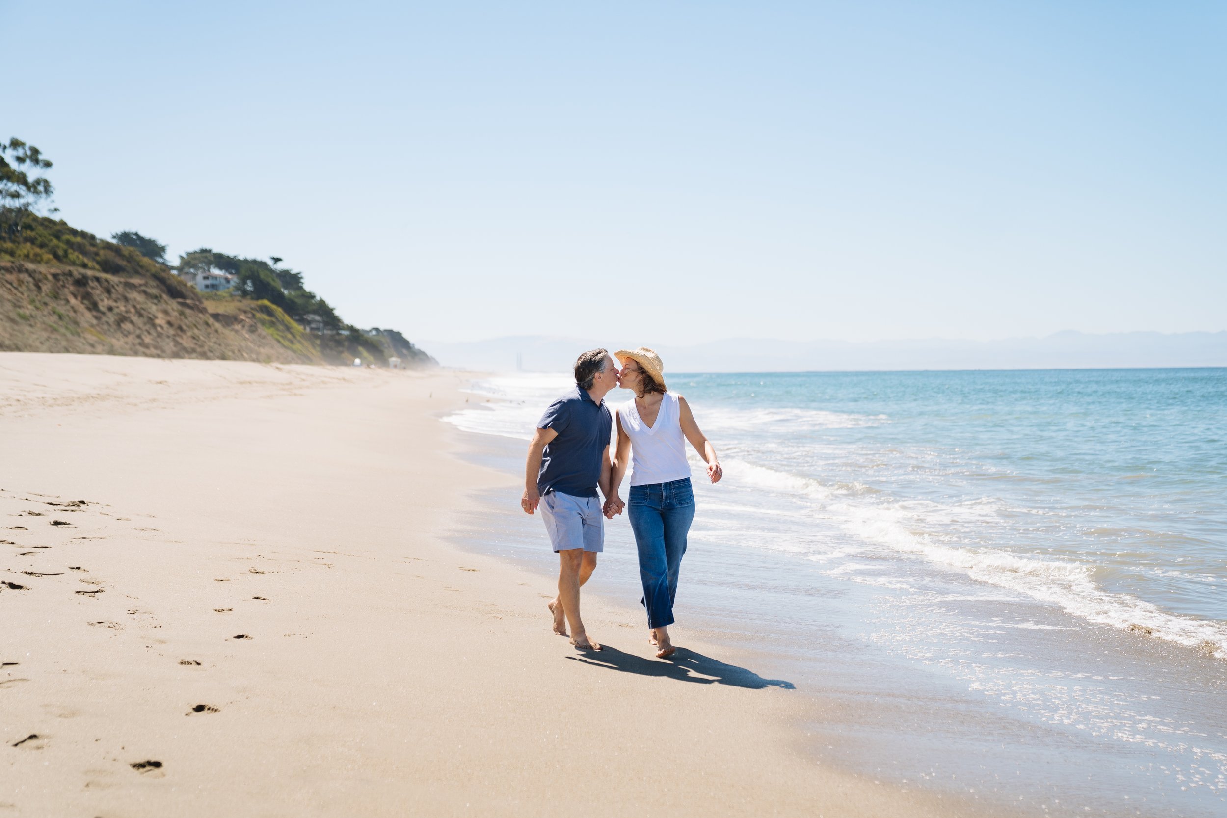 A couple walking hand in hand along the shoreline of a sandy beach, kissing, with blue ocean water and a clear sky in the background.