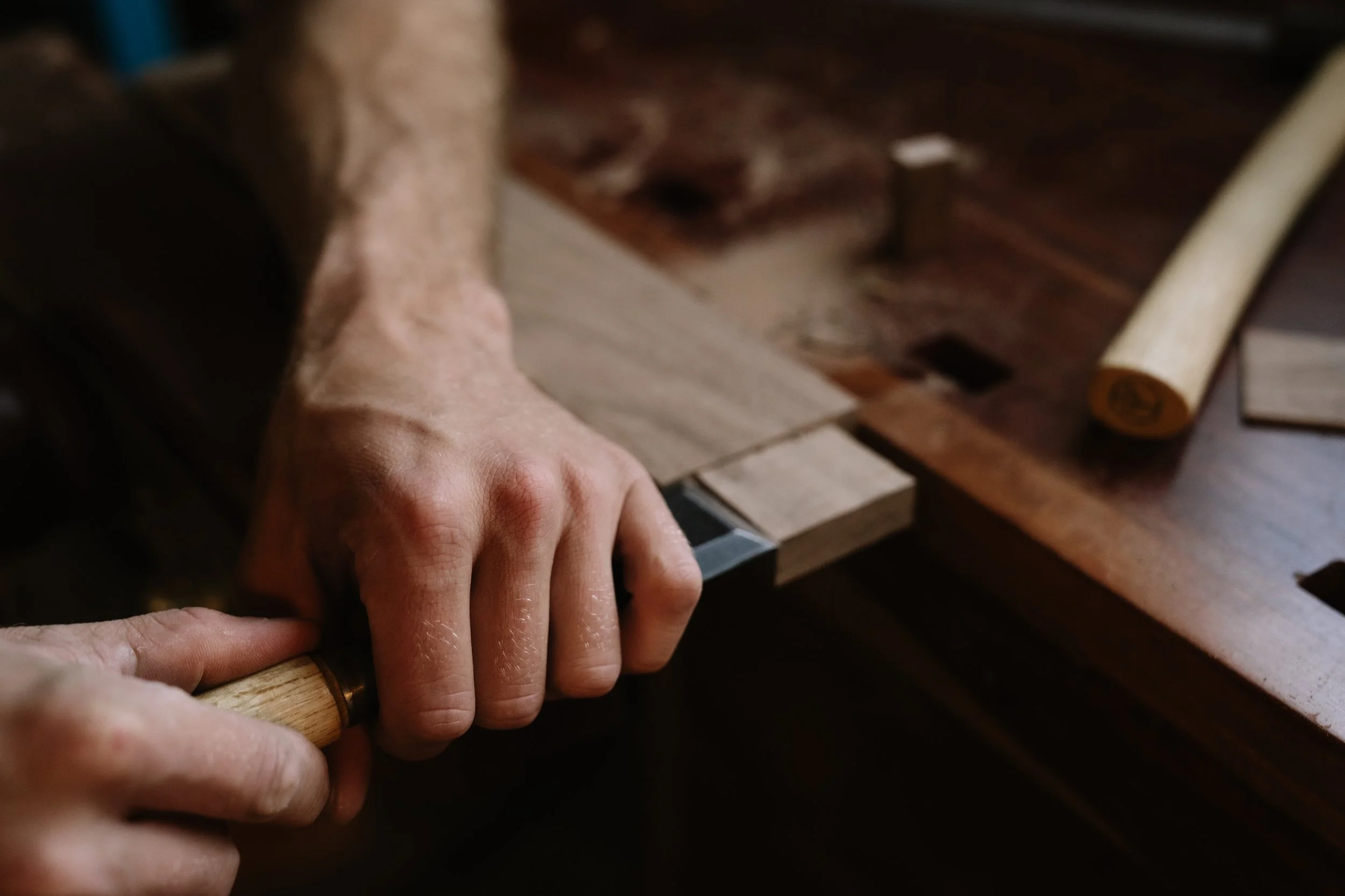 Close-up of a person hand using a hammer to hammer a nail into a piece of wood on a workbench.