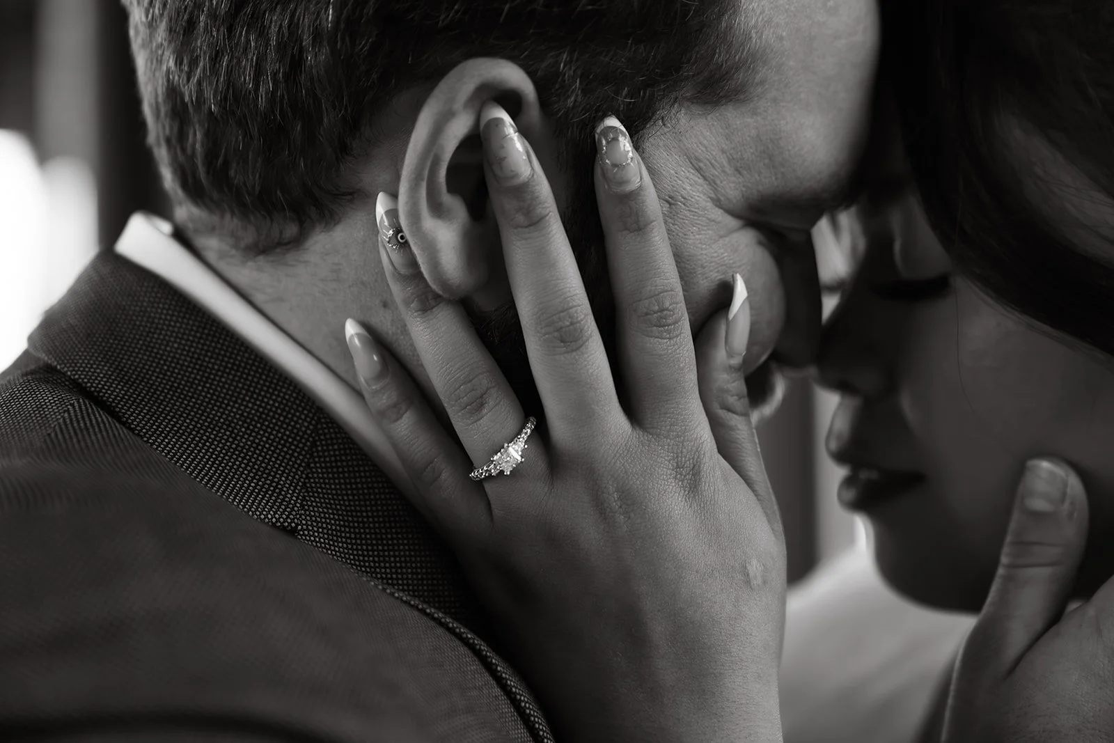 Close-up black-and-white photograph of a couple touching foreheads. The woman has her hand on the man's face, showcasing rings and manicured nails. The woman appears to be embracing the man tenderly.