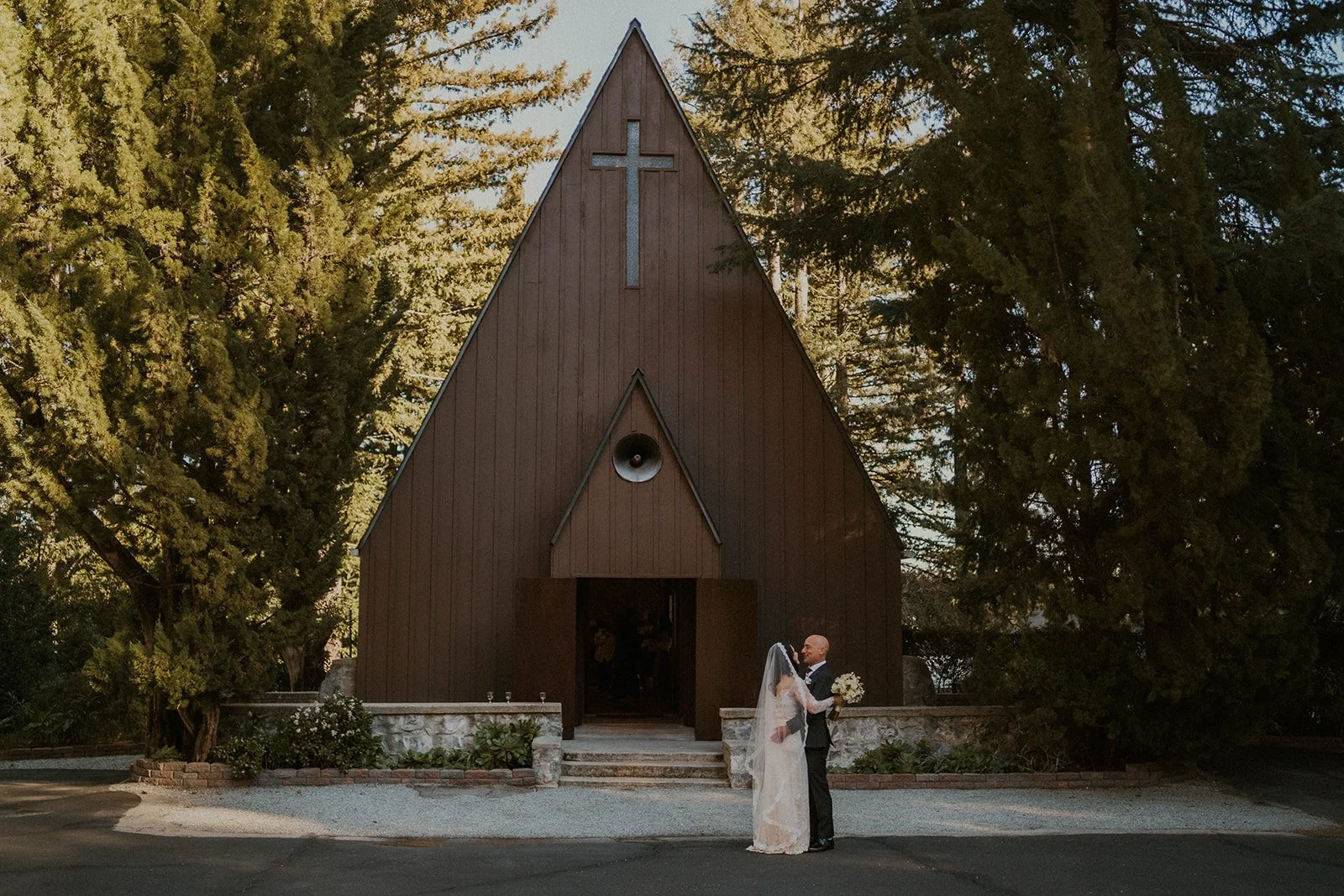 A bride and groom standing close in front of a small wooden church with a cross on the front, surrounded by tall trees, during a wedding.