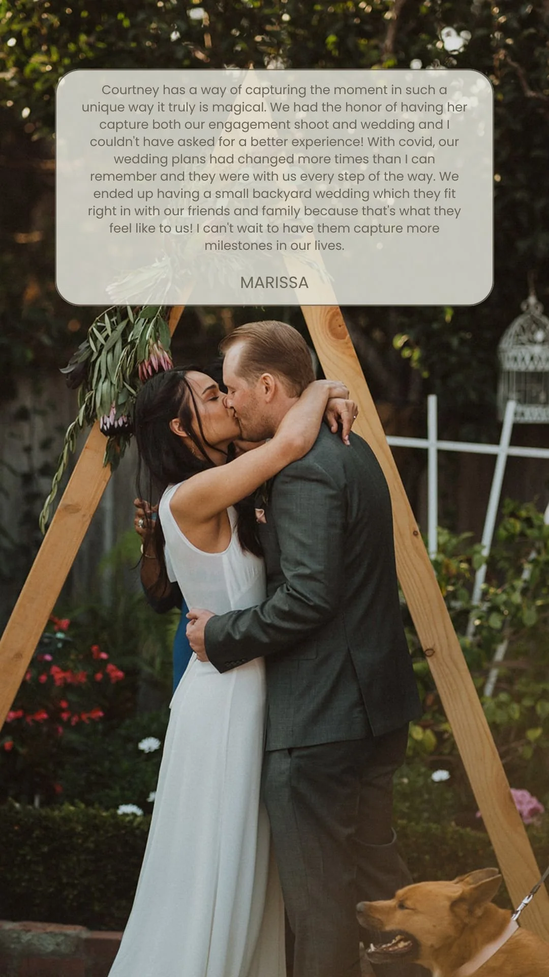 A couple kissing in front of a wooden wedding arch decorated with flowers at an outdoor wedding ceremony. A brown dog is sitting at their feet.