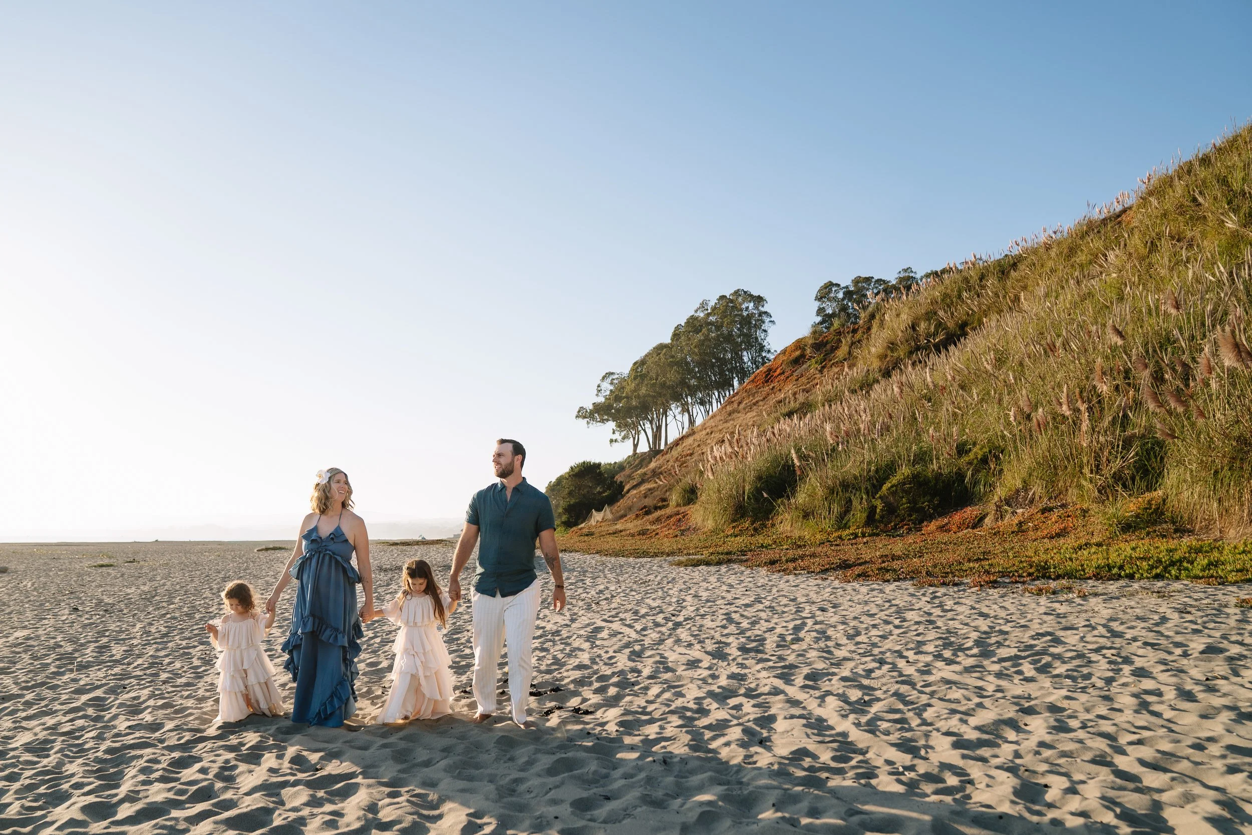 A family of four walking on a sandy beach near a grassy cliff under a clear blue sky. The mother and father hold hands with two young girls.