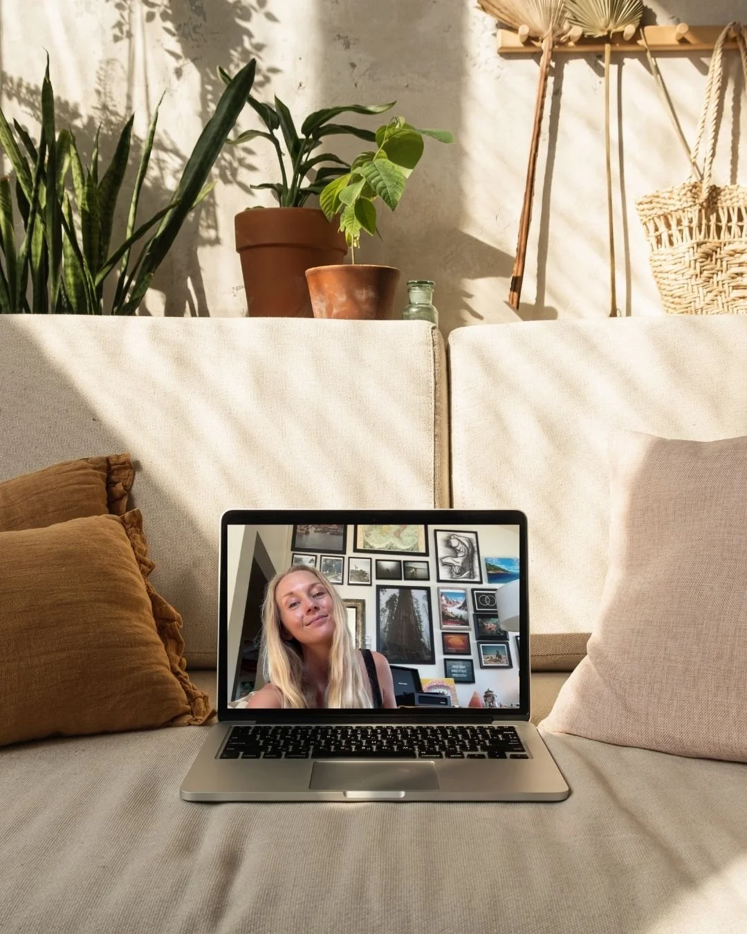 A laptop on a beige sofa showing a woman with long blonde hair, smiling, in front of a gallery wall of framed pictures.