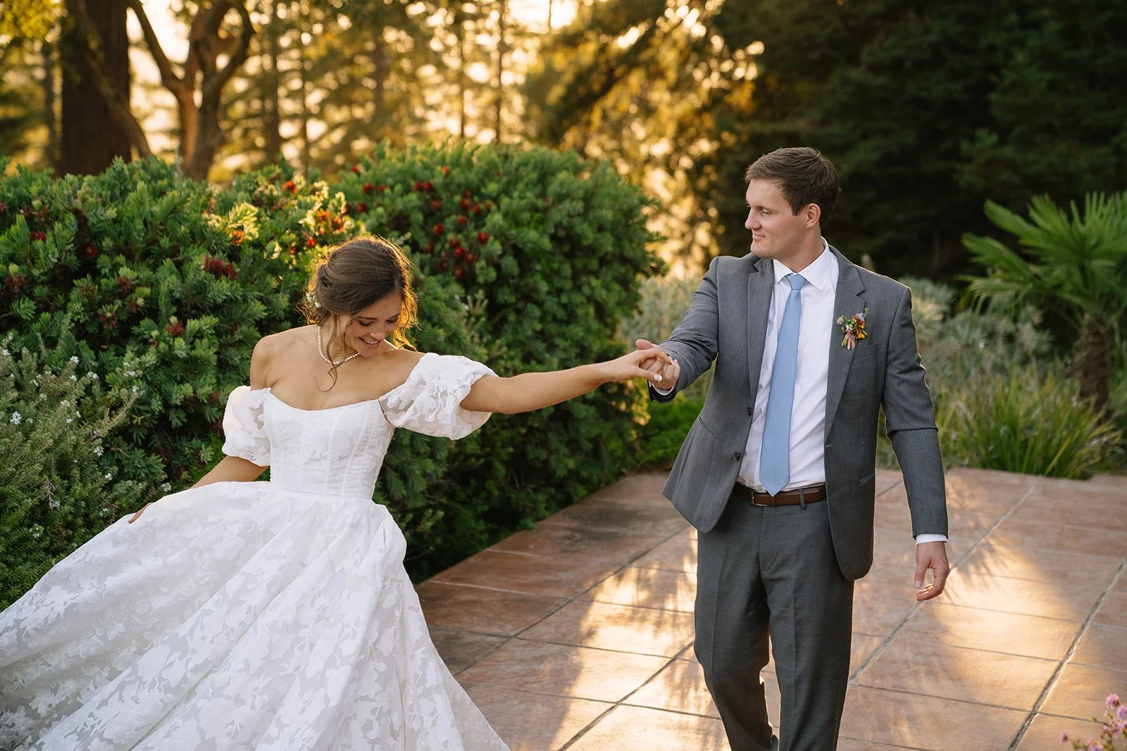 A bride and groom dancing outdoors during a wedding ceremony at sunset, with lush greenery and flowering bushes in the background.
