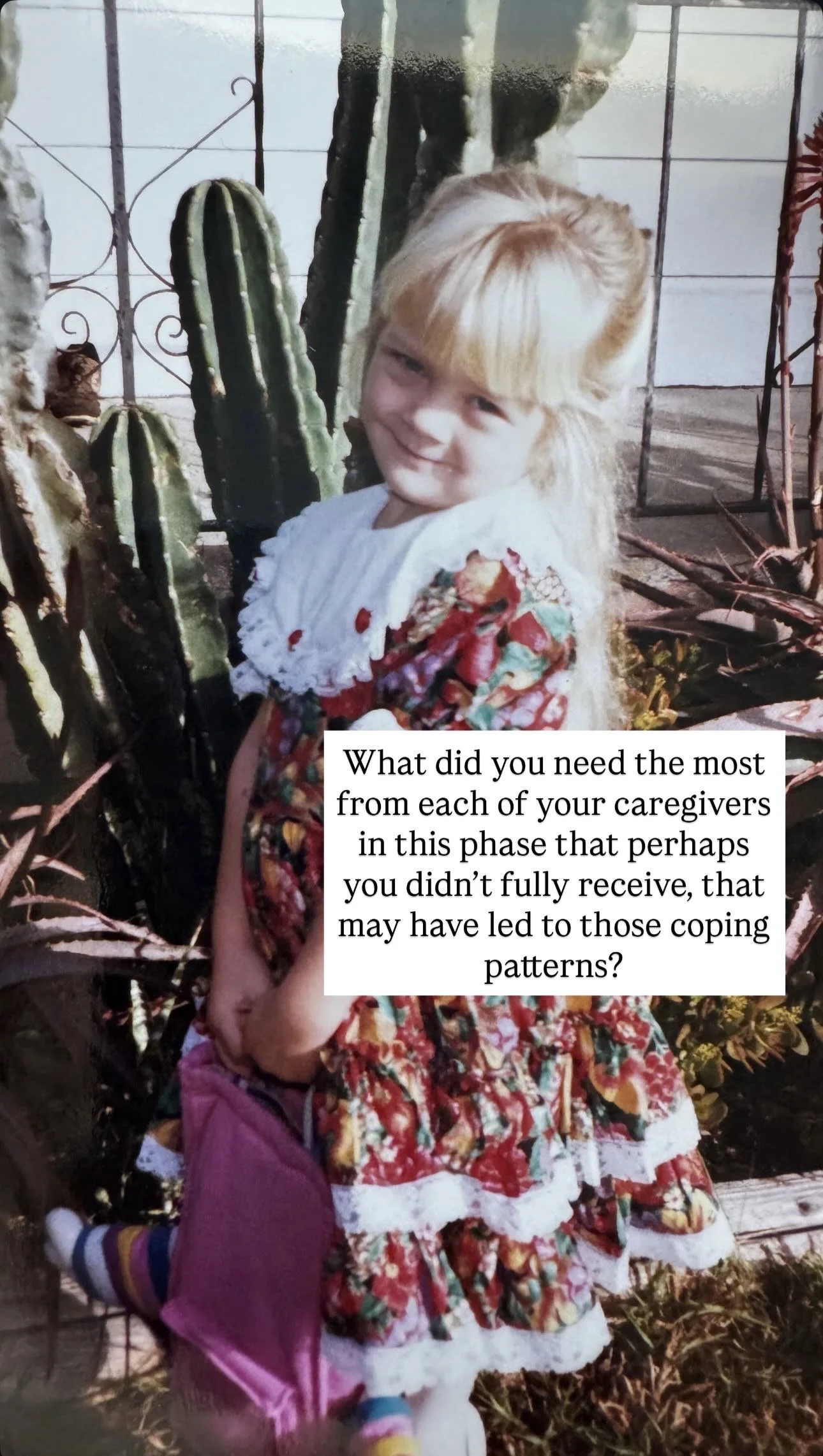 A young girl with blonde hair in a bun and wearing a colorful floral dress, standing outdoors next to cacti and garden plants, smiling at the camera.