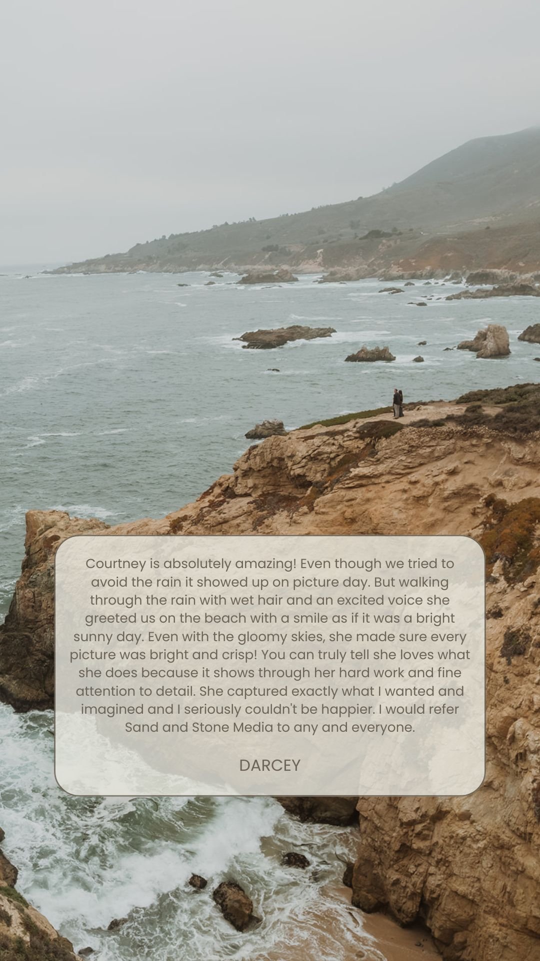 A person standing on a rocky cliff overlooking a rainy coastline with water crashing against rocks, under a cloudy sky.