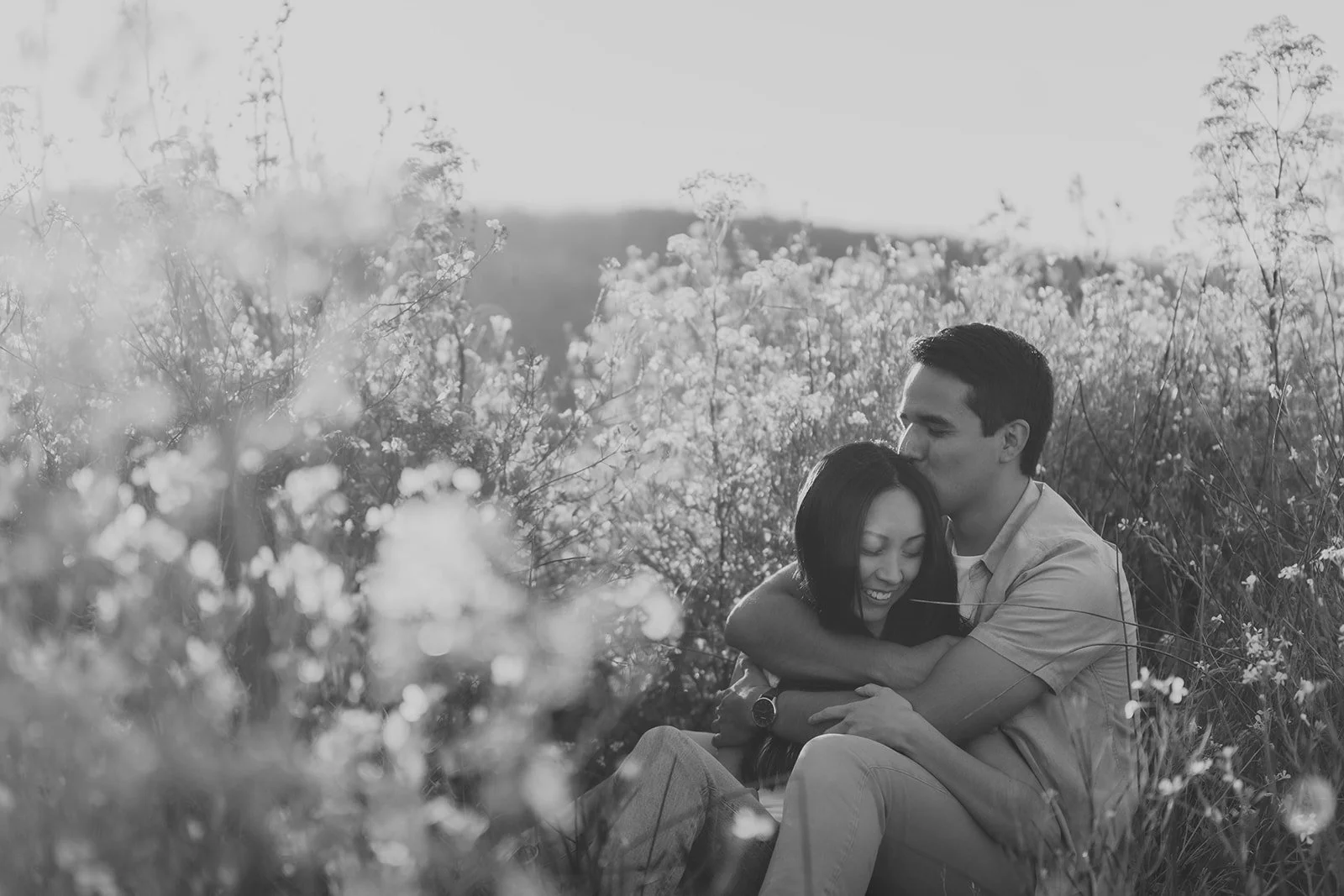 A black and white photo of a young couple sitting in a field of flowers, embracing and sharing a kiss.