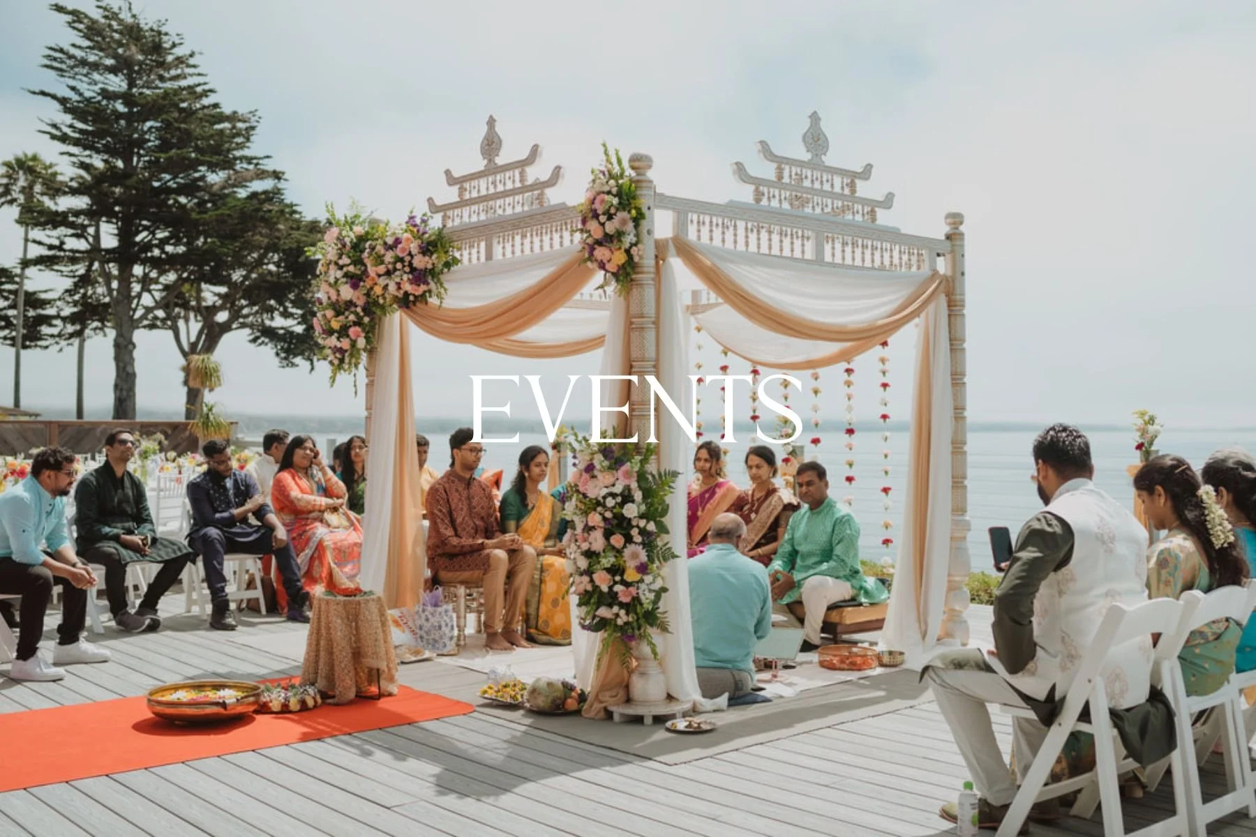 A traditional outdoor Indian wedding ceremony on a deck overlooking water, with floral decorations, a canopy, and guests seated on white chairs.