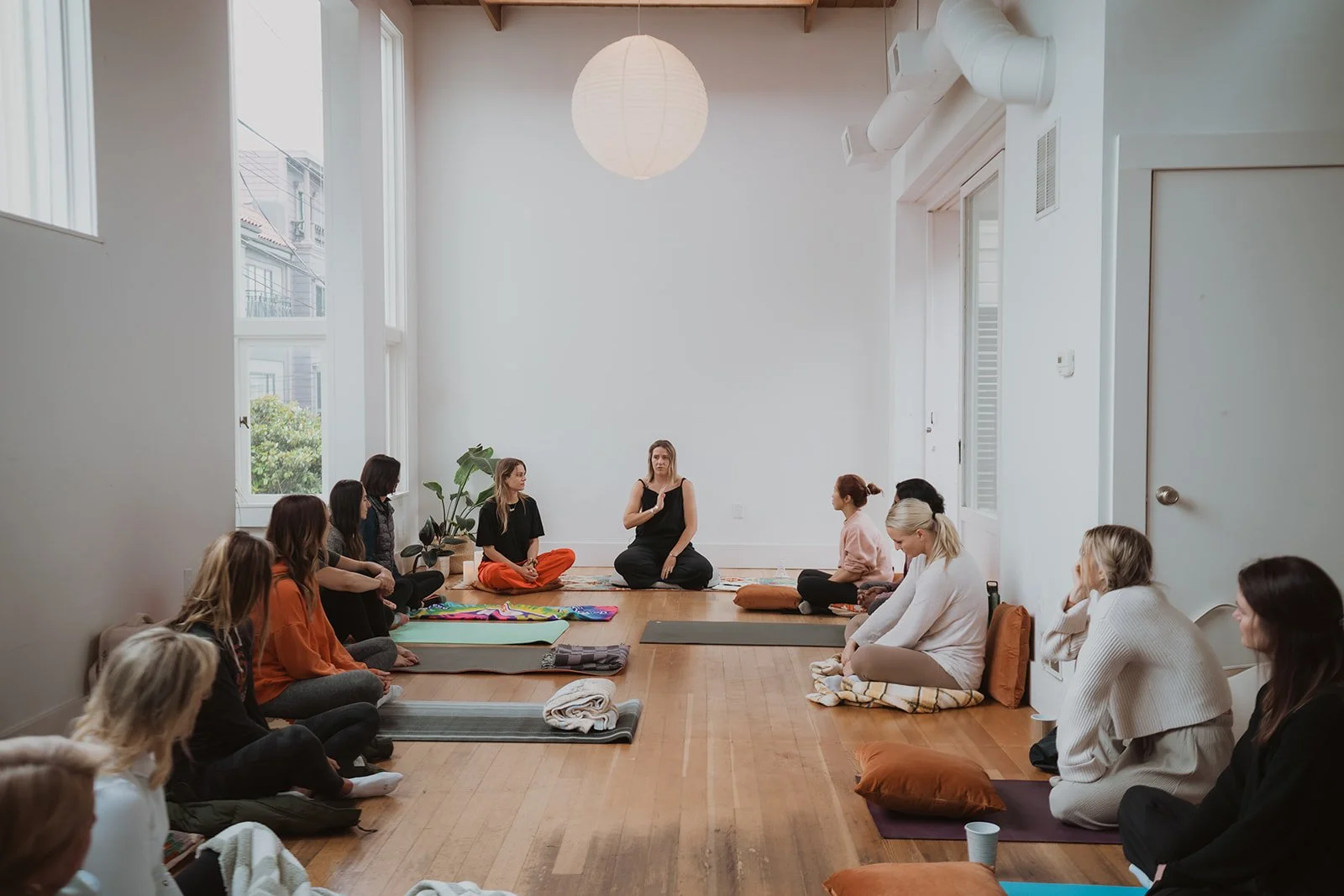 A group of people sitting cross-legged on yoga mats and cushions in a bright, minimalist room, participating in a meditation or yoga session led by an instructor sitting at the front.