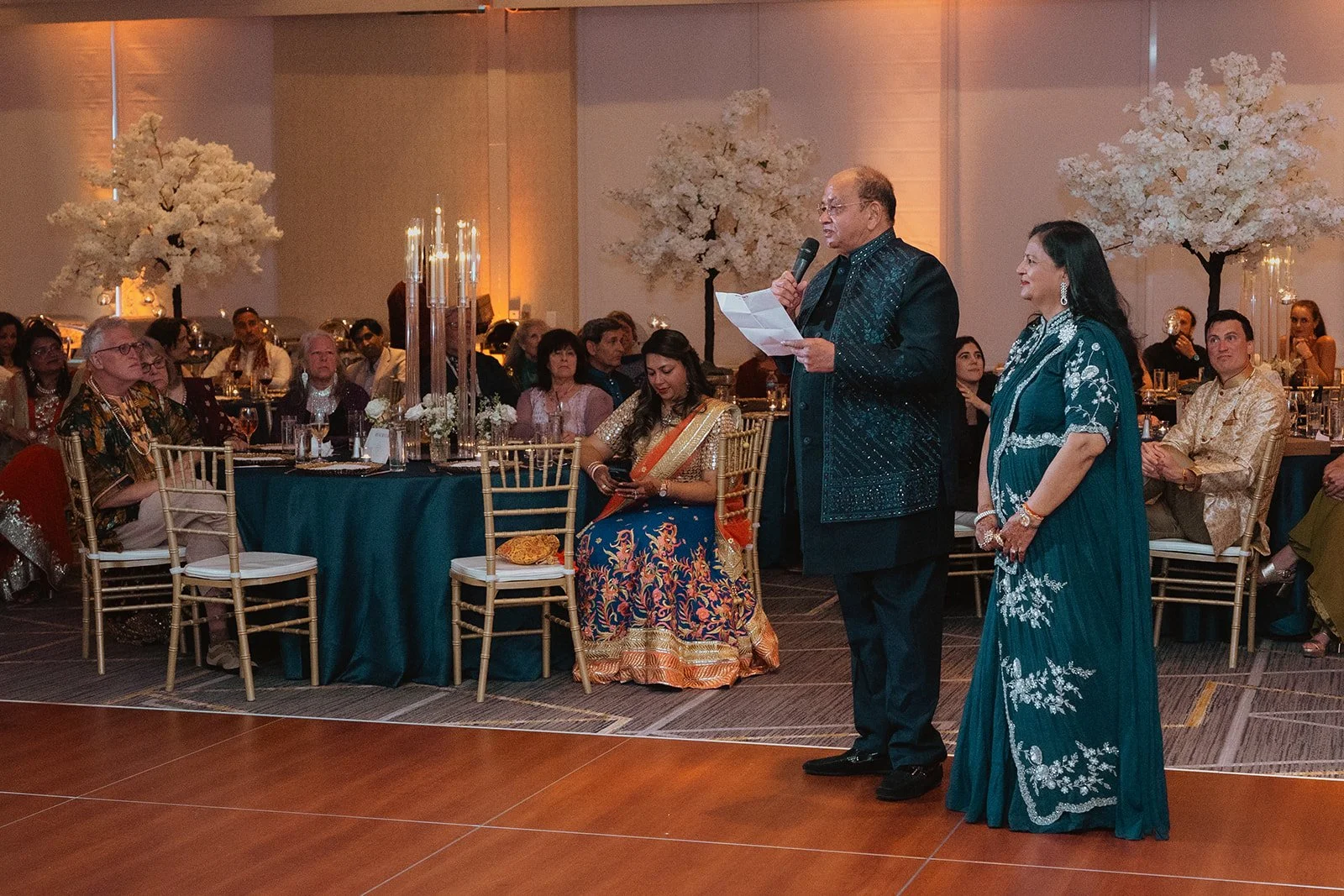 Man and woman speaking on stage at formal event, surrounded by seated guests, decorated with white trees, candles, and elegant table settings.