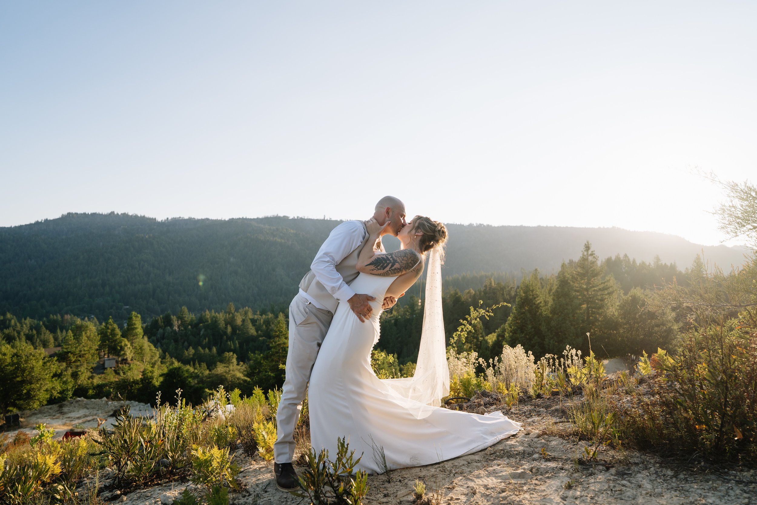 A couple in wedding attire sharing a kiss outdoors during sunset, with mountains and trees in the background.