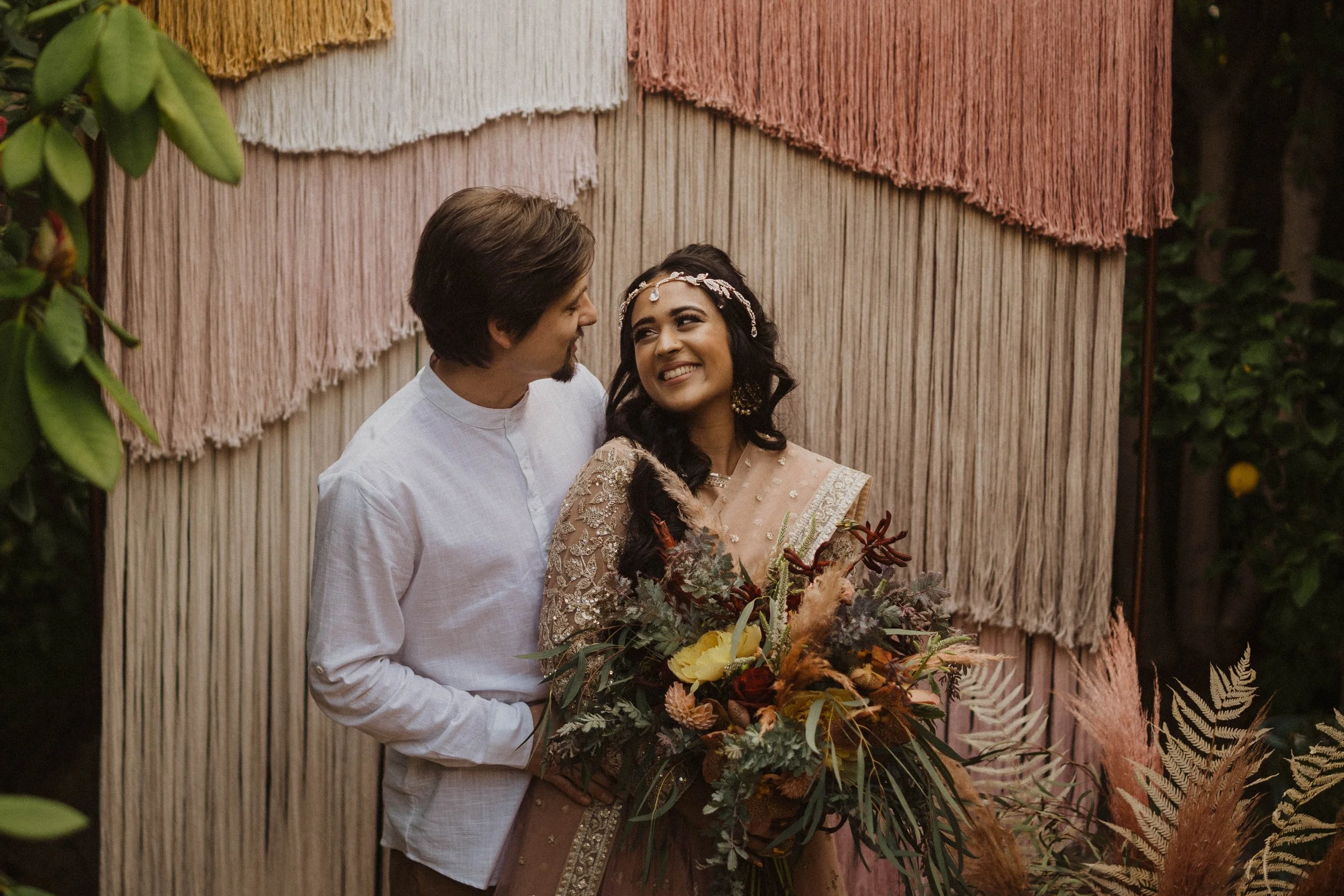 A couple at a wedding, with the man wearing a white shirt and the woman in a gold dress holding a bouquet of flowers, standing in front of a textured backdrop with hanging fabric.