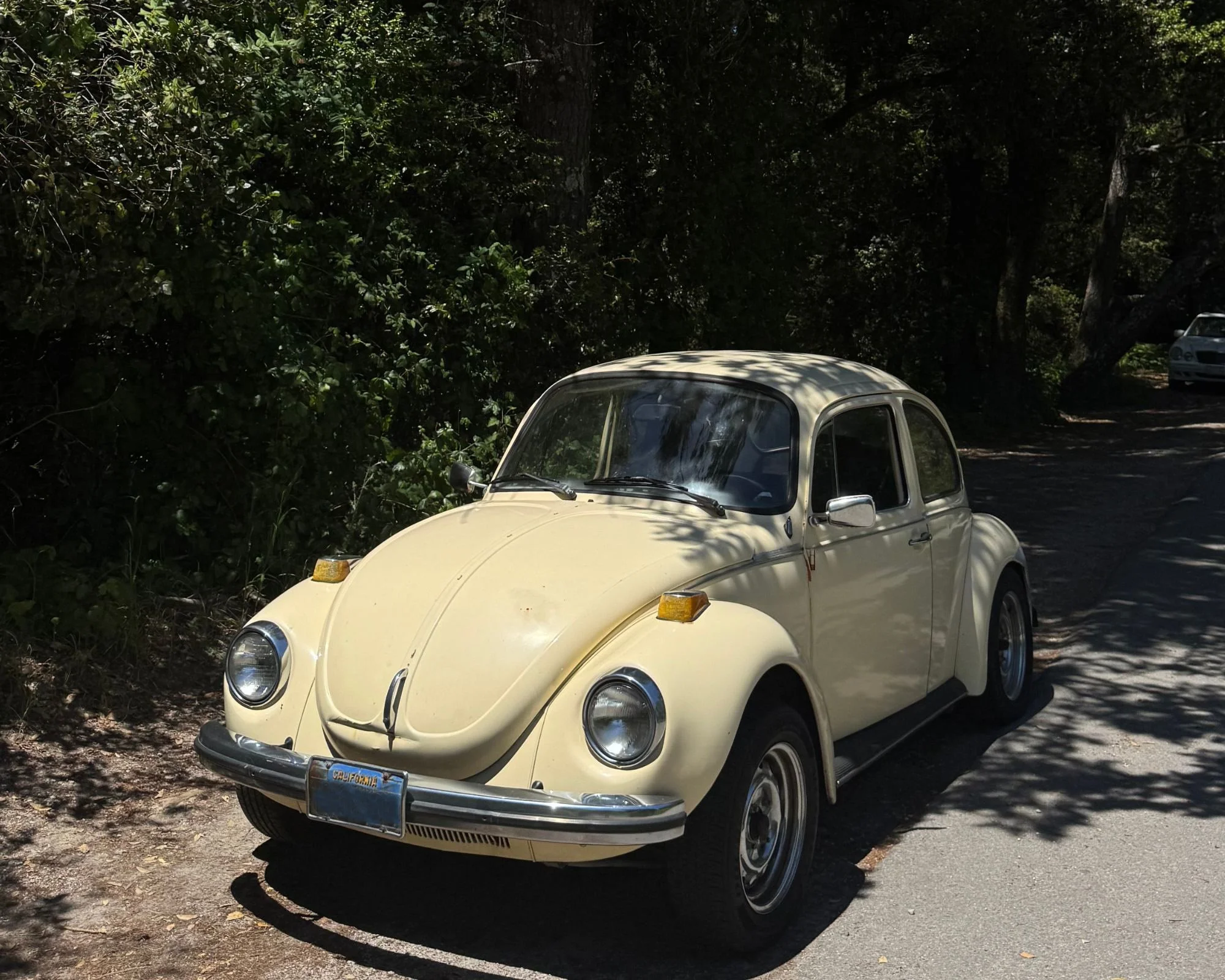 A vintage yellow Volkswagen Beetle parked on the side of a shaded road with trees in the background.