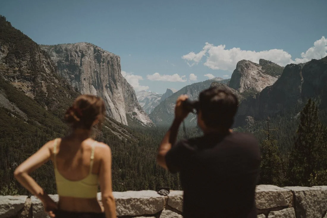 Two people, a woman and a man, standing on a stone wall overlooking a mountain landscape, with the man taking a photo of the view.