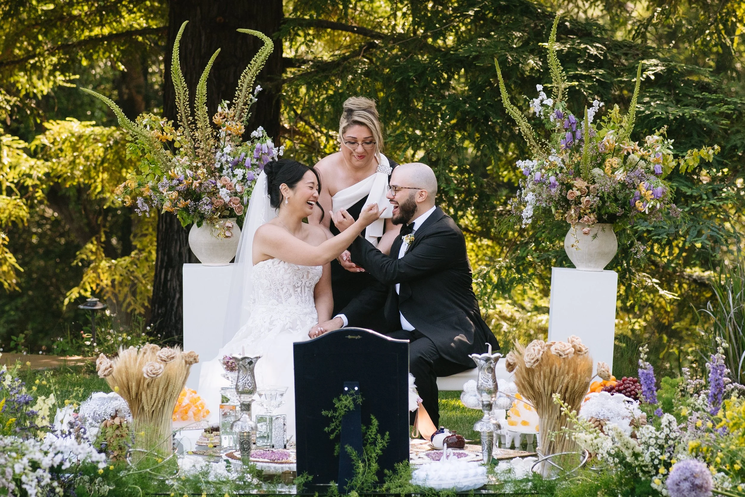 A wedding ceremony with a couple, a woman in a white wedding dress and a man in a black suit, sitting outdoors at a decorated table, smiling and touching each other's faces. An officiant is standing behind them, overseeing the ceremony. The scene is set with large floral arrangements in vases, greenery, and a variety of flowers, with trees in the background.