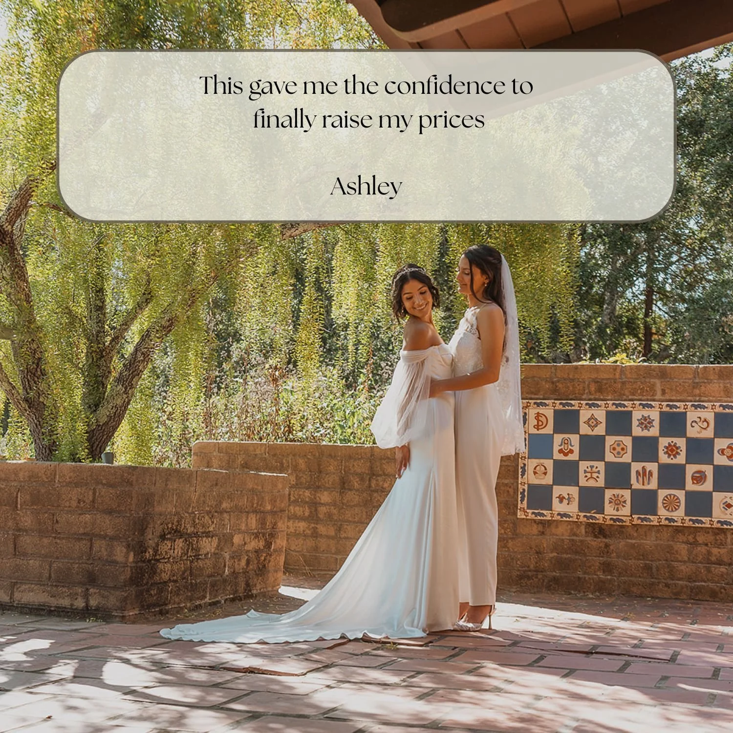 Two women dressed in white wedding gowns standing close together outdoors, smiling. One woman is wearing a veil. The background features trees, a brick wall, and patterned tiles.
