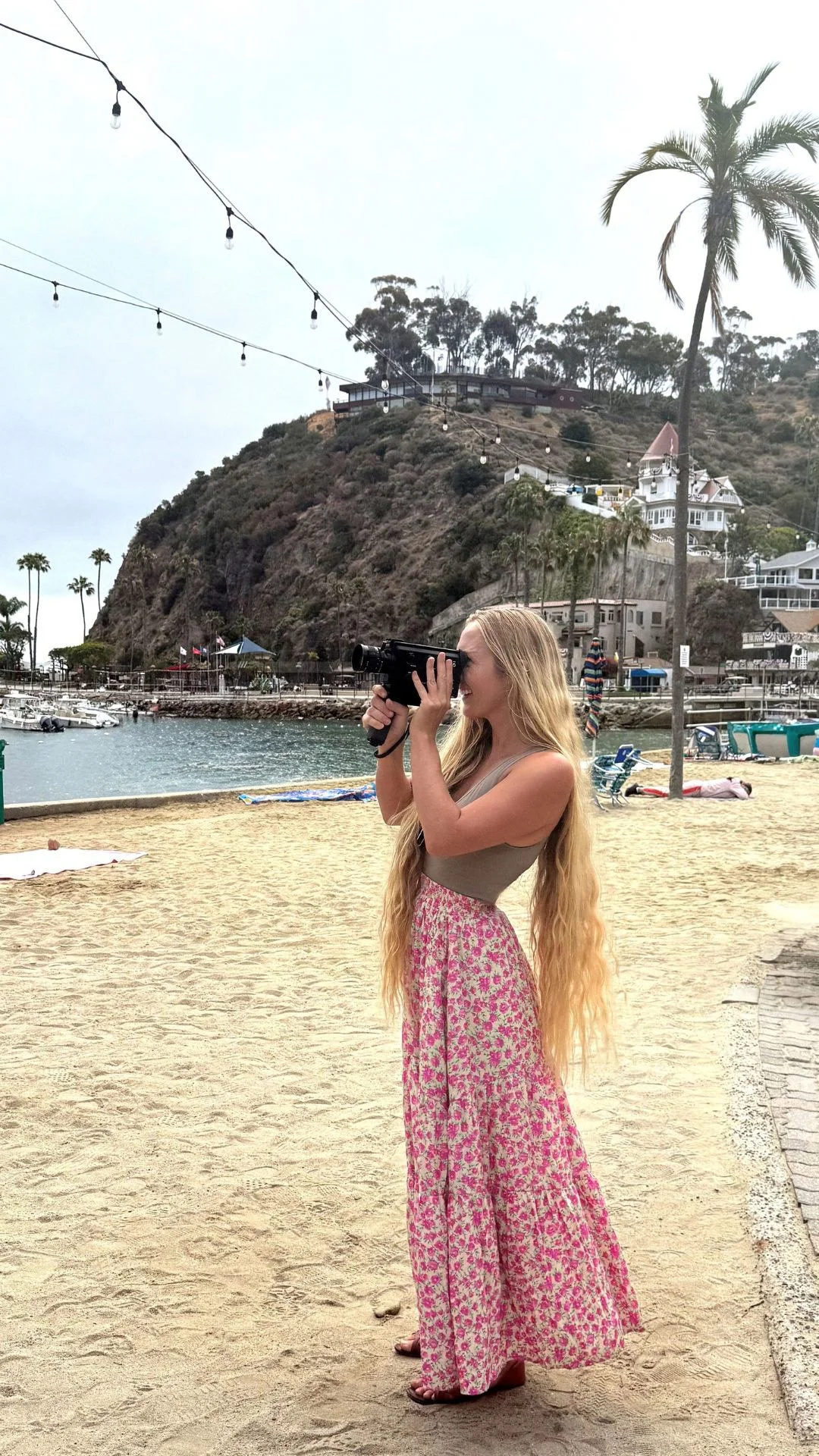A woman with long blonde hair wearing a beige top and a pink floral skirt is standing on a sandy beach, taking a photo with a vintage camera. In the background, there are palm trees, boats in the water, and houses on a hillside, with string lights hanging overhead.