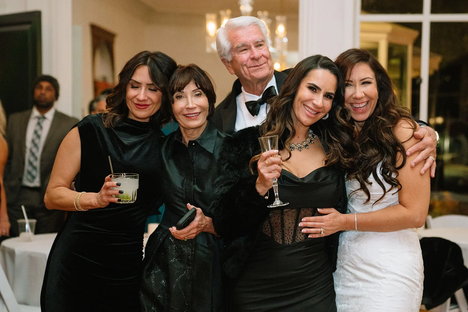 Group of five people dressed in formal attire, celebrating at a party or event with drinks in hand.