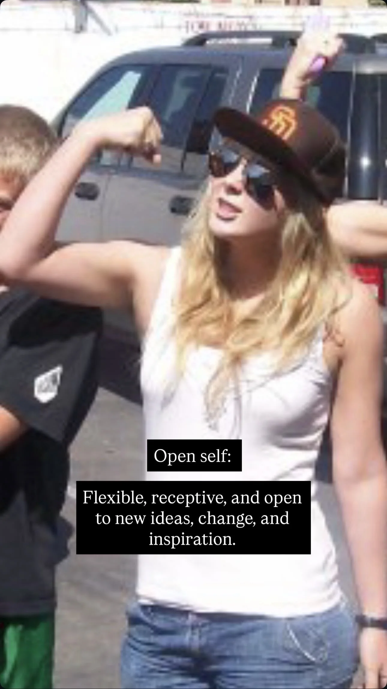 A woman wearing sunglasses and a San Francisco Giants cap flexes her bicep while standing outdoors near parked cars, with a boy partly visible beside her.