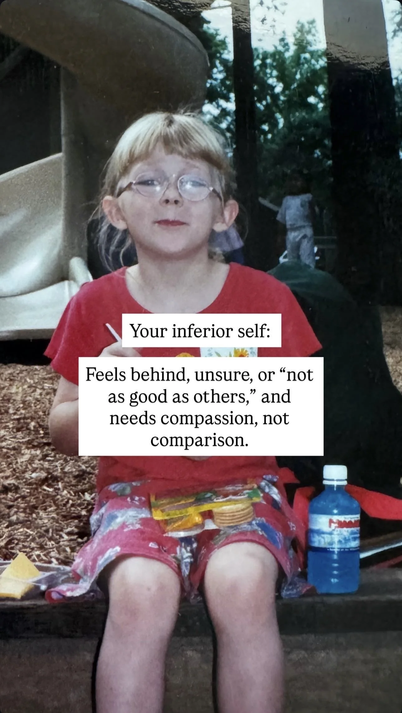 A young girl with glasses, sitting outdoors, holding a snack. There is a large water bottle and snacks in front of her. The background shows trees and a reflection of a person.