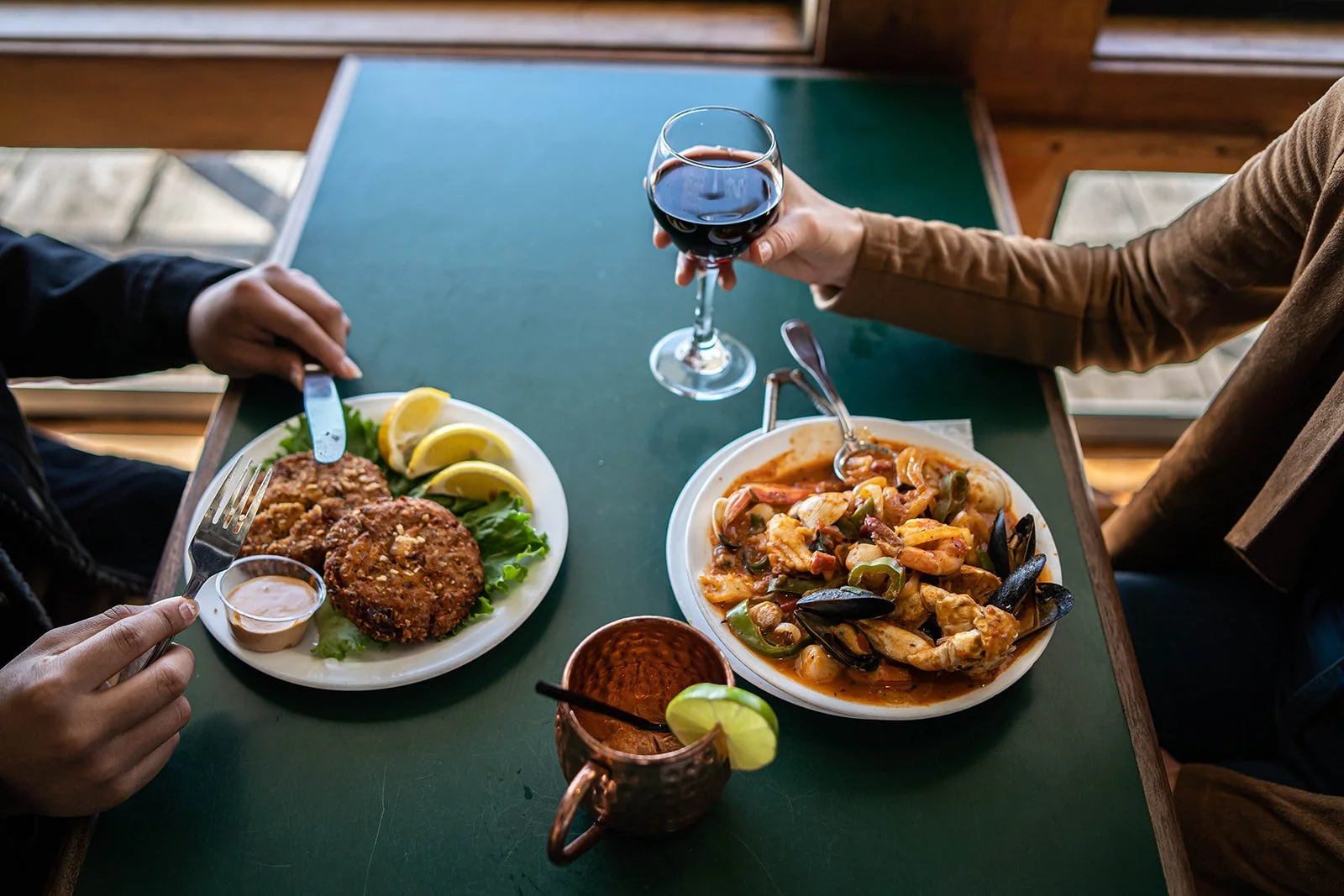 Two people sharing a meal at a restaurant, with plates of fried chicken with lemon wedges and a seafood pasta dish, and drinks including red wine and a cocktail with lime.