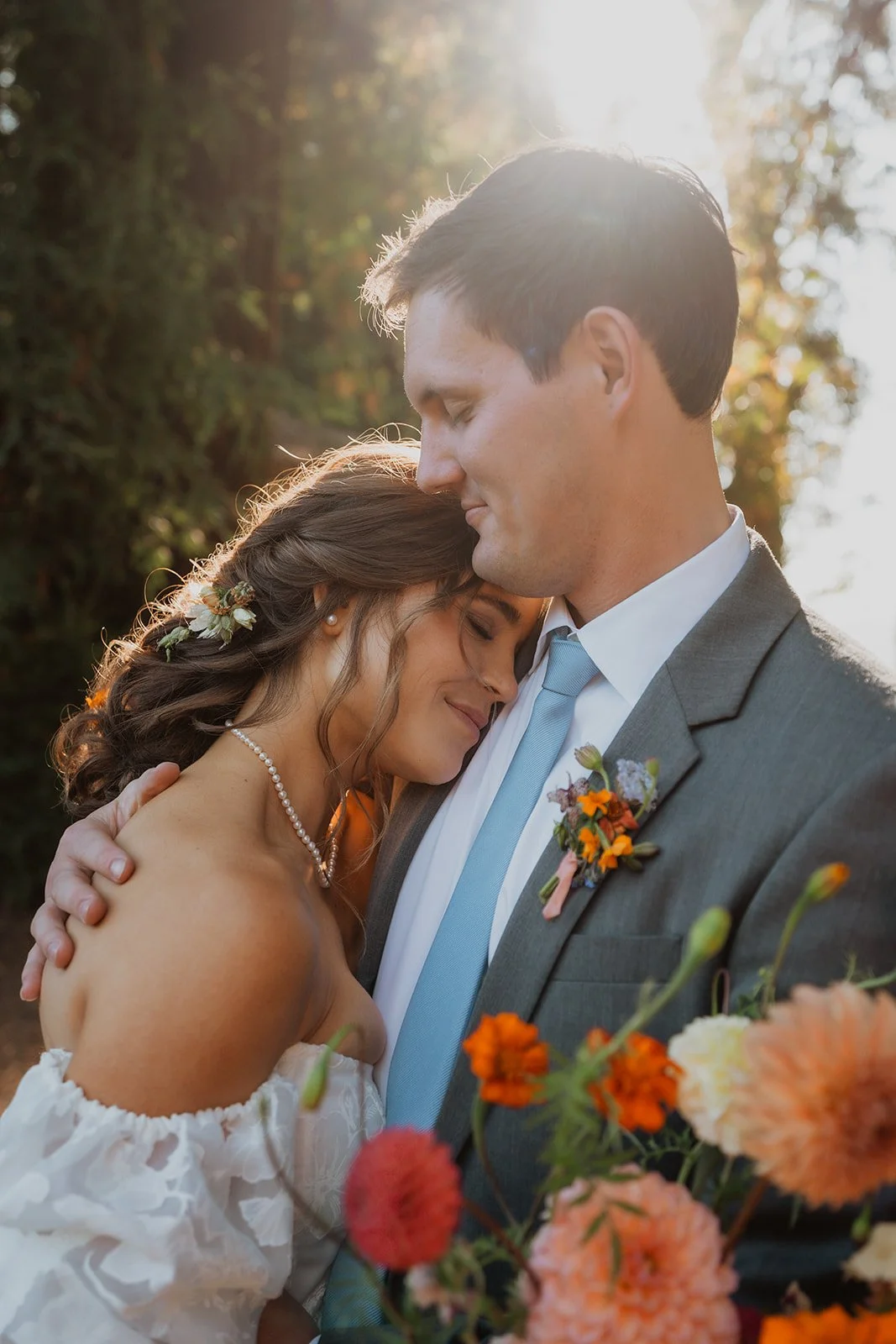 A bride and groom embrace outdoors during sunset, with trees in the background and flowers in the foreground.