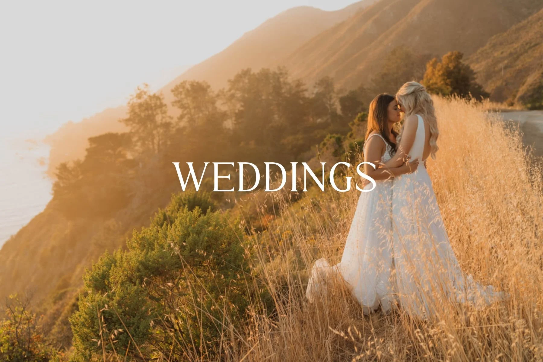 Two women in wedding dresses sharing an intimate moment outdoors during sunset, with mountainous landscape and tall grasses in the background.