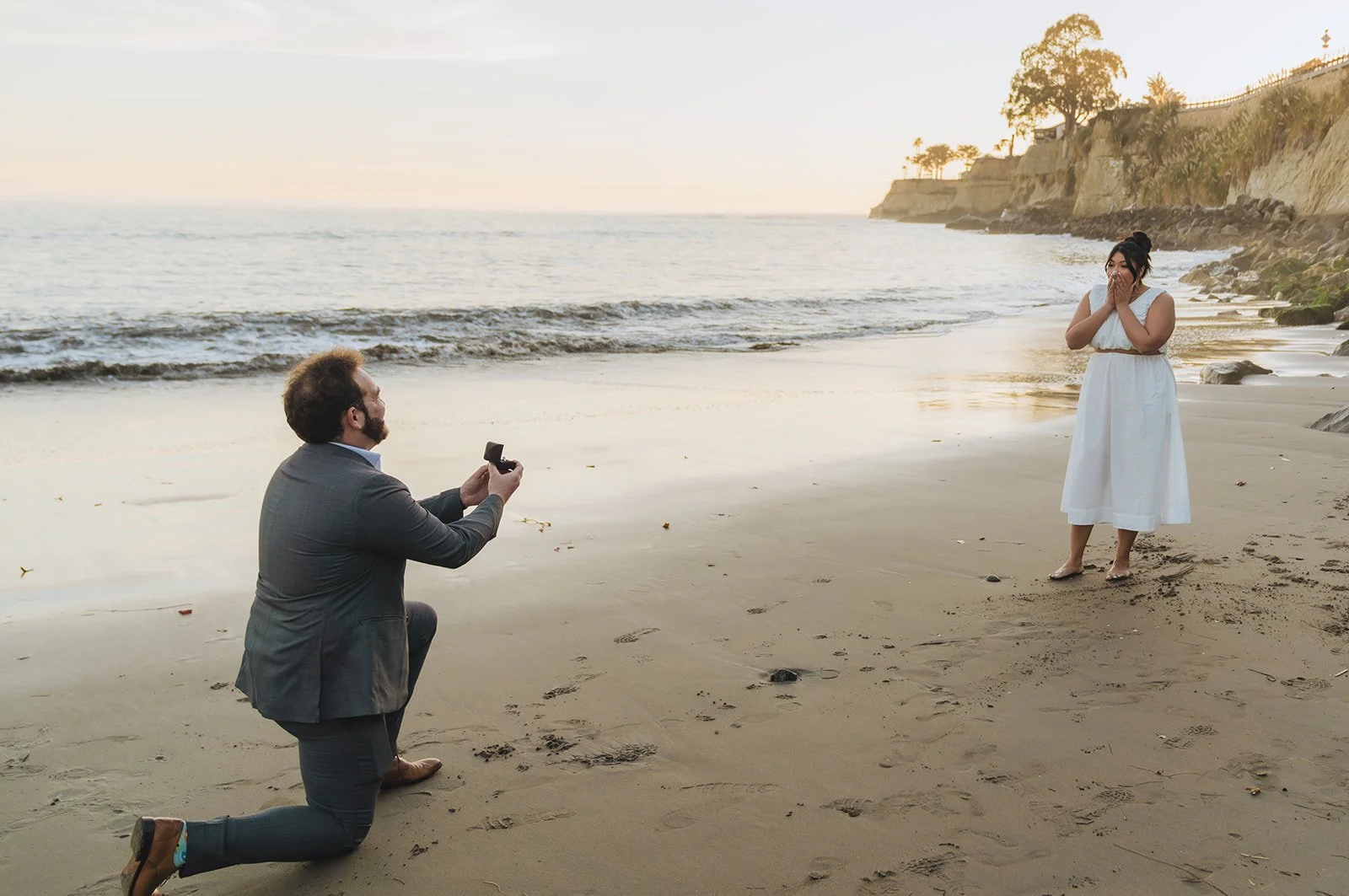 A man kneeling on the beach proposing to a woman who is wearing a white dress and covering her mouth in surprise with her hands.