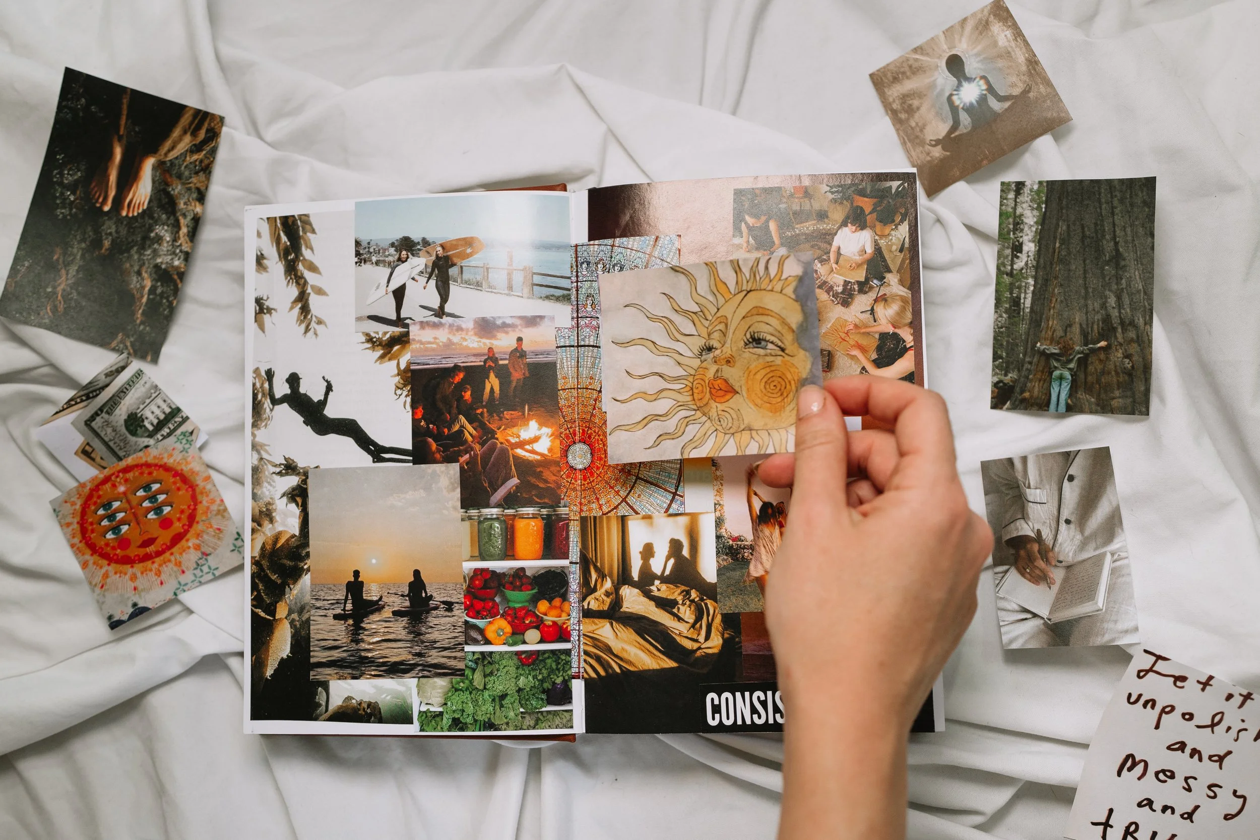 A collage of various photographs and artwork in a magazine, surrounded by loose photos, a small drawing of a sun with a face, colorful jars, and a handwritten note on a white fabric surface.