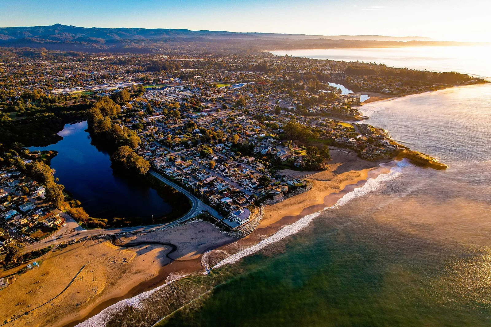 Aerial view of a coastal residential area with houses lining the shore, a beach, and a body of water, during sunset.