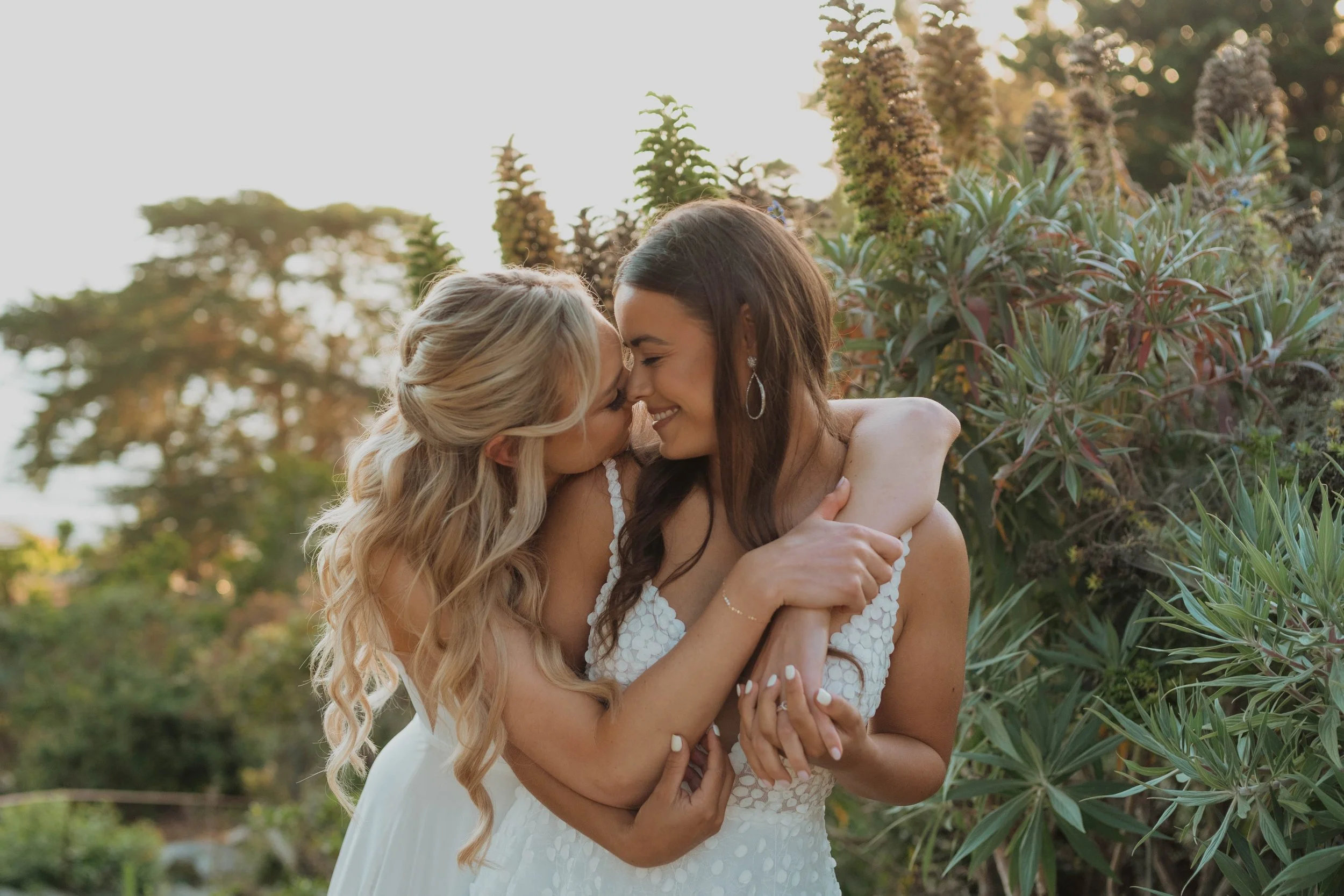 Two women, one with blonde hair and the other with dark brown hair, sharing a joyful embrace outdoors during sunset, surrounded by lush greenery.
