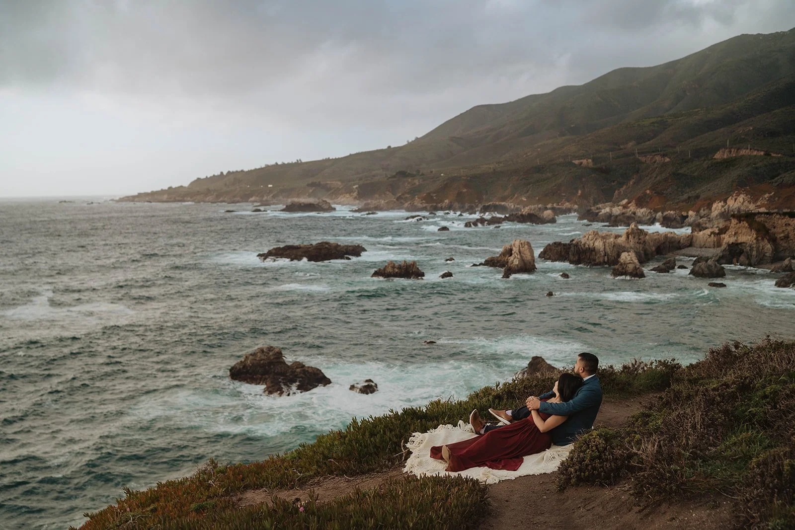 A couple sitting on a blanket on a cliff overlooking the ocean and rocky shoreline with hills in the background on a cloudy day.